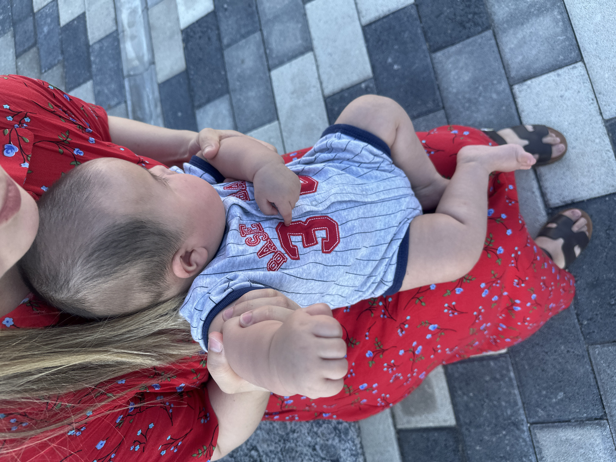The image shows a baby being held by an adult. The baby is wearing a striped onesie with red lettering that appears to say "BASE" and a number 3. The adult is wearing a red dress with a floral pattern. The scene appears to be outdoors, with a stone-tiled ground visible. The lighting suggests it is daytime, and the weather is likely pleasant. The overall mood is one of tenderness and connection between the adult and the baby. While the specific location is stated as Manas, Kyrgyzstan, there are no specific visual cues in the image to confirm this. The focus is on the personal moment between the two individuals.