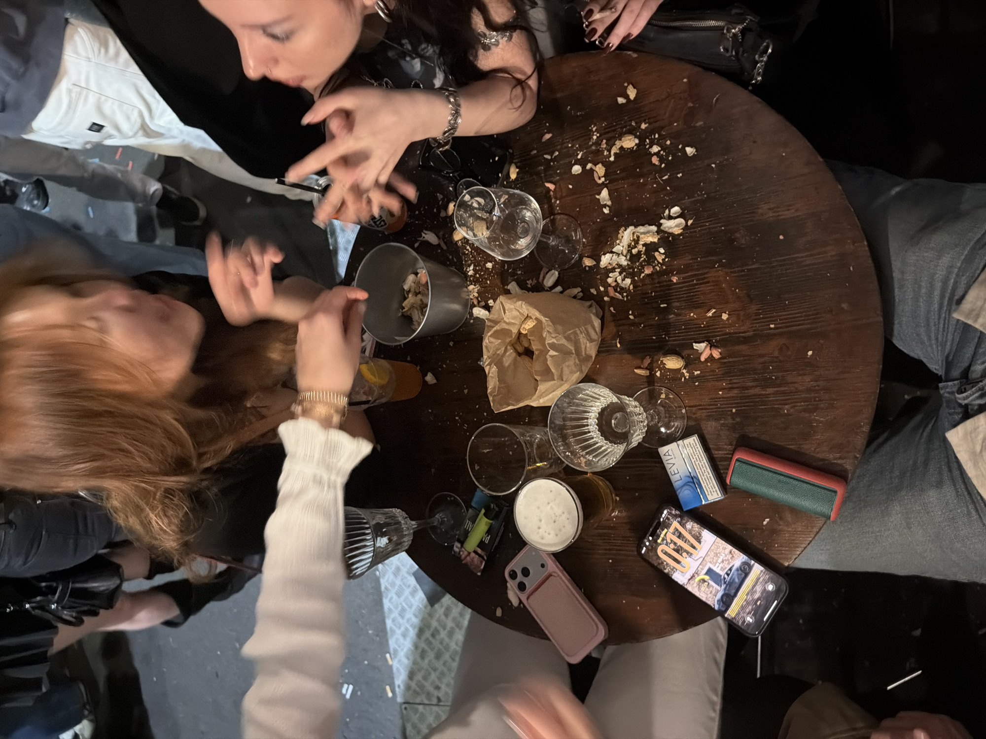 The image depicts a circular wooden table in a dimly lit environment, possibly a bar or pub. Several individuals are seated around the table. The table surface is covered with peanut shells and discarded food debris. There are multiple empty and partially filled glasses, a metal bucket containing peanut shells, a paper bag with peanuts, a phone with a car on the screen, a pack of cigarettes, a wallet and a pint of beer. The location of the photo being in Milan, Italy is NOT clear from visual clues within the image.