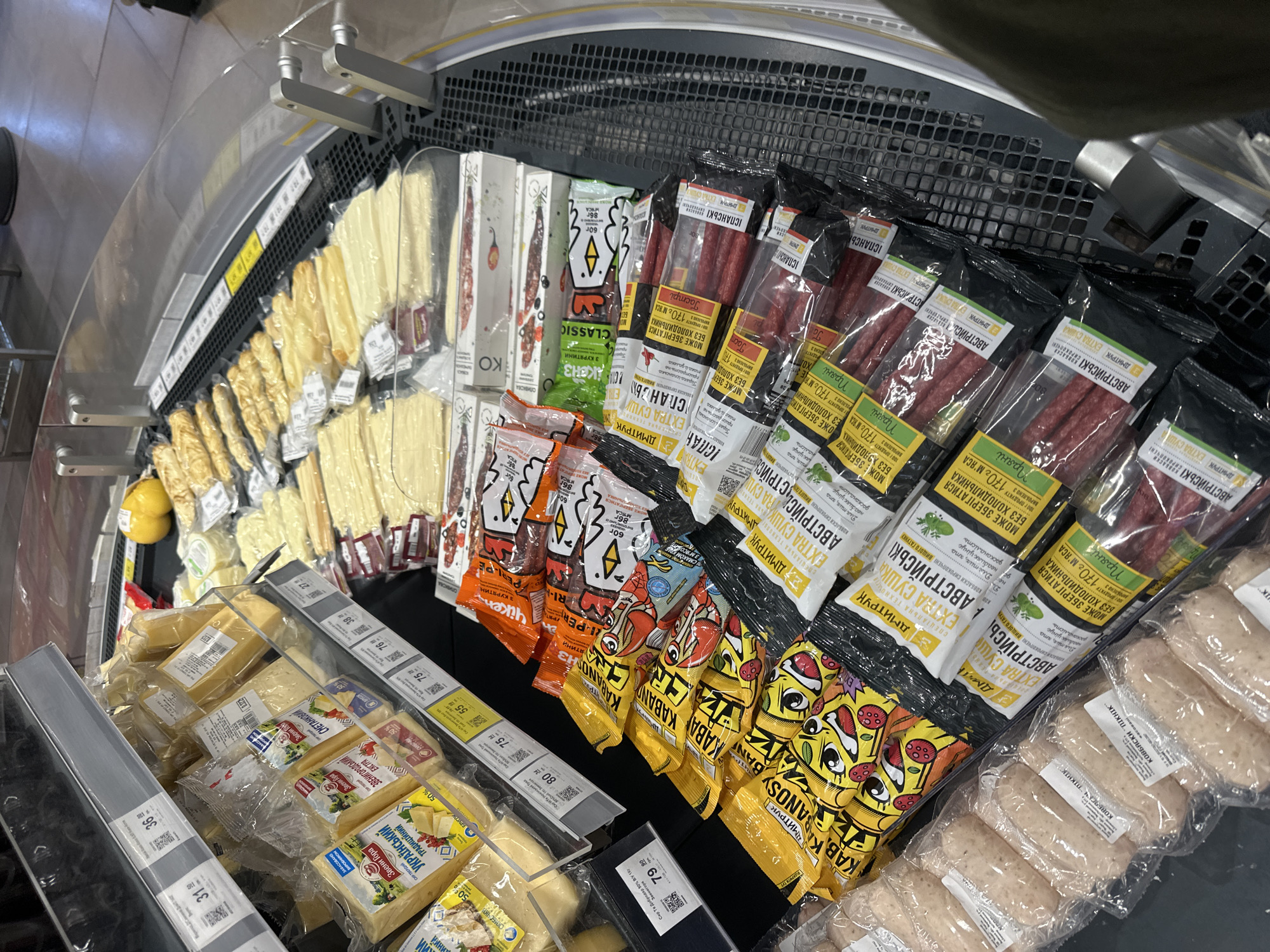 A mid-shot, eye-level photograph displays a refrigerated section of a supermarket in Ternopil, Ukraine, likely an aisle within the store, with a curved glass barrier in the foreground. The display is filled with a variety of packaged cheeses and sausages. The cheese products are visible in the upper left of the frame, with labels featuring Ukrainian text and images of dairy farms. Below the cheese, an array of packaged snack sausages are neatly arranged. These sausages come in bright, cartoonish packaging, predominantly yellow and orange, with humorous depictions of chickens and fruits. Further down, darker packaged sausages with more mature-themed designs are stacked. The lighting appears to be overhead, typical of a retail environment, illuminating the products clearly. The overall scene suggests a typical grocery shopping experience, with products organized for customer selection.