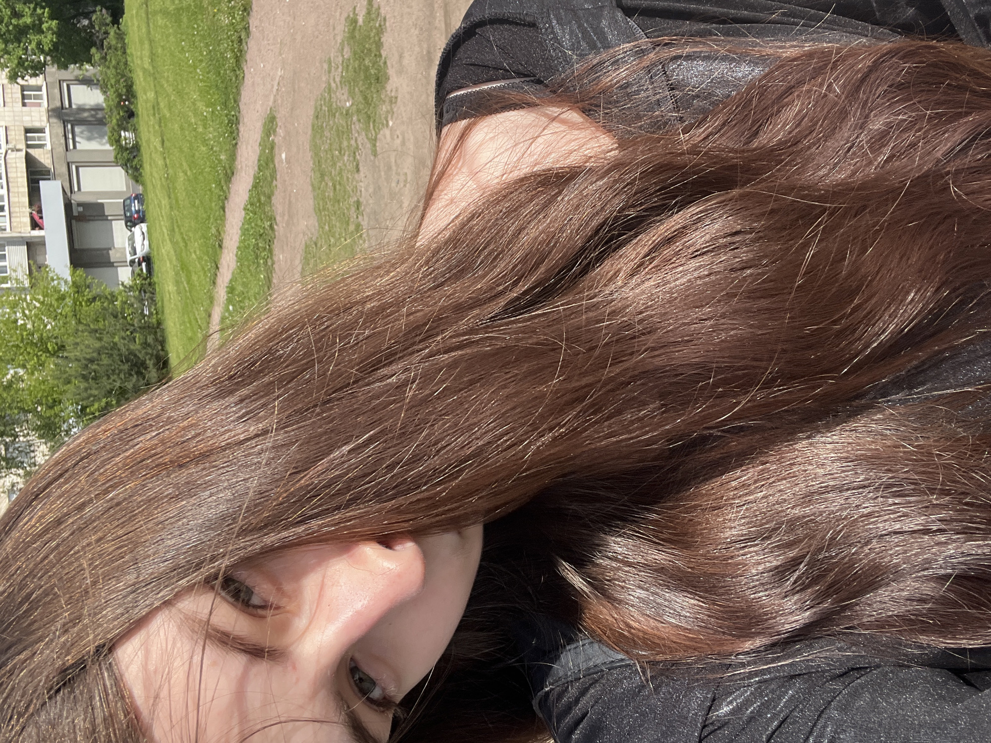The image is a close-up portrait, oriented on its side, featuring a person with long, flowing dark brown hair. Sunlight illuminates the hair, revealing warm highlights and a glossy texture. Only a portion of the person's face is visible in the lower-left corner of the frame, including one eye with a dark iris, part of the nose, and a cheek, suggesting a casual, relaxed moment. The person wears a dark, possibly black, shimmering or textured garment on their upper body.
The background, appearing on the left side of the rotated image, reveals an outdoor urban scene. A wide area of green grass is cut by a winding dirt path. In the mid-distance, multi-story residential or commercial buildings with numerous windows and a relatively simple facade are visible. Two cars, one dark and one white, are parked near these structures, surrounded by scattered trees and foliage.
The strong, direct sunlight and clear shadows indicate a bright, sunny day. The architectural style of the buildings suggests an urban environment, consistent with the provided context of Saint Petersburg, Russia. No specific activity is clearly discernible, nor is any text visible.