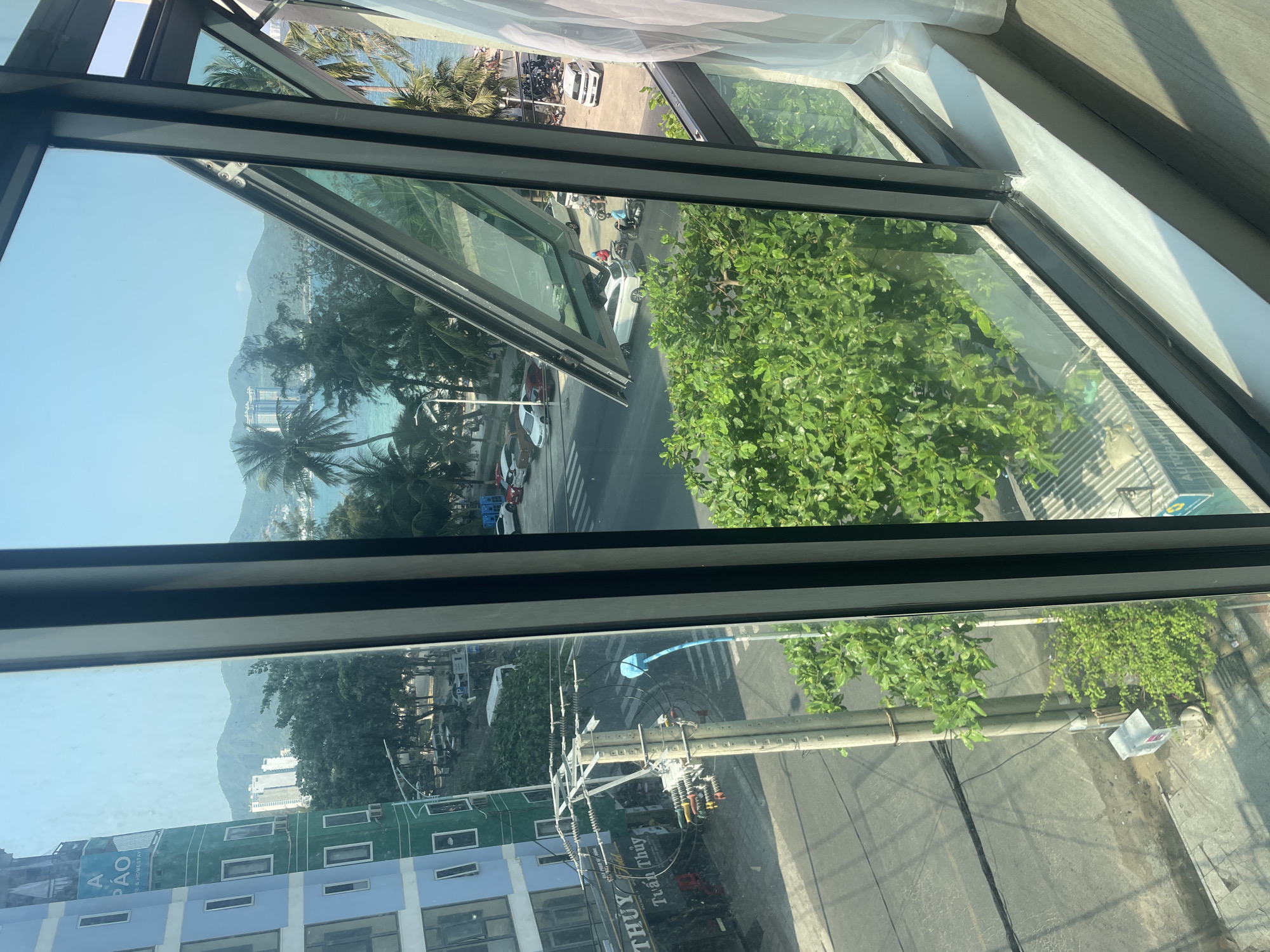 The scene is viewed through a modern window, partially open, offering a view of Bắc Nha Trang, Vietnam. The foreground consists of the window frame itself and a glimpse of a white curtain. The midground shows a street scene with a mix of vehicles, including cars and motorbikes, parked along the road. Palm trees line the beach, and further in the background, there's a view of mountains and the ocean. The lower part of the window frame shows buildings with blue and green facades. A utility pole with wires and transformers is also visible, running alongside the street. There is no visible interactions or people in the scene.