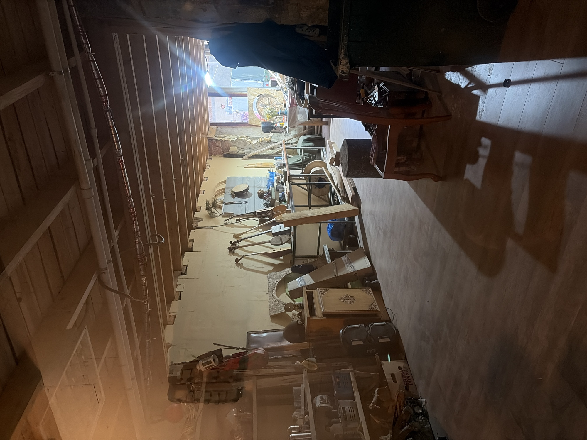 The image shows the interior of a workshop or studio, possibly in Il'inskiy Pogost, Russia. The space appears to be dimly lit, with natural light streaming in from a window or opening at the far end. The ceiling is made of exposed wooden beams and planks, with visible pipes and electrical conduits running across.

The room is cluttered with various objects, suggesting it's an active workspace. On the left side, there are shelves filled with containers, bottles, and other miscellaneous items. To the right of the shelves, a wooden cabinet stands against the wall, adorned with decorative carvings. Several musical instruments, possibly stringed instruments like lutes or dulcimers, are propped against the wall and displayed on a rack.

In the foreground and mid-ground, there are chairs, wooden planks, and a tree stump acting as a stand or work surface. A dark blue garment is draped over a chair in the mid-right portion of the image. The floor is made of wooden planks. The back wall has a rough, textured appearance, with exposed brickwork in some areas. In the far background, through an opening, a person can be seen standing near what appears to be a large circular art piece. The overall impression is that of a functional, albeit somewhat unorganized, creative space.