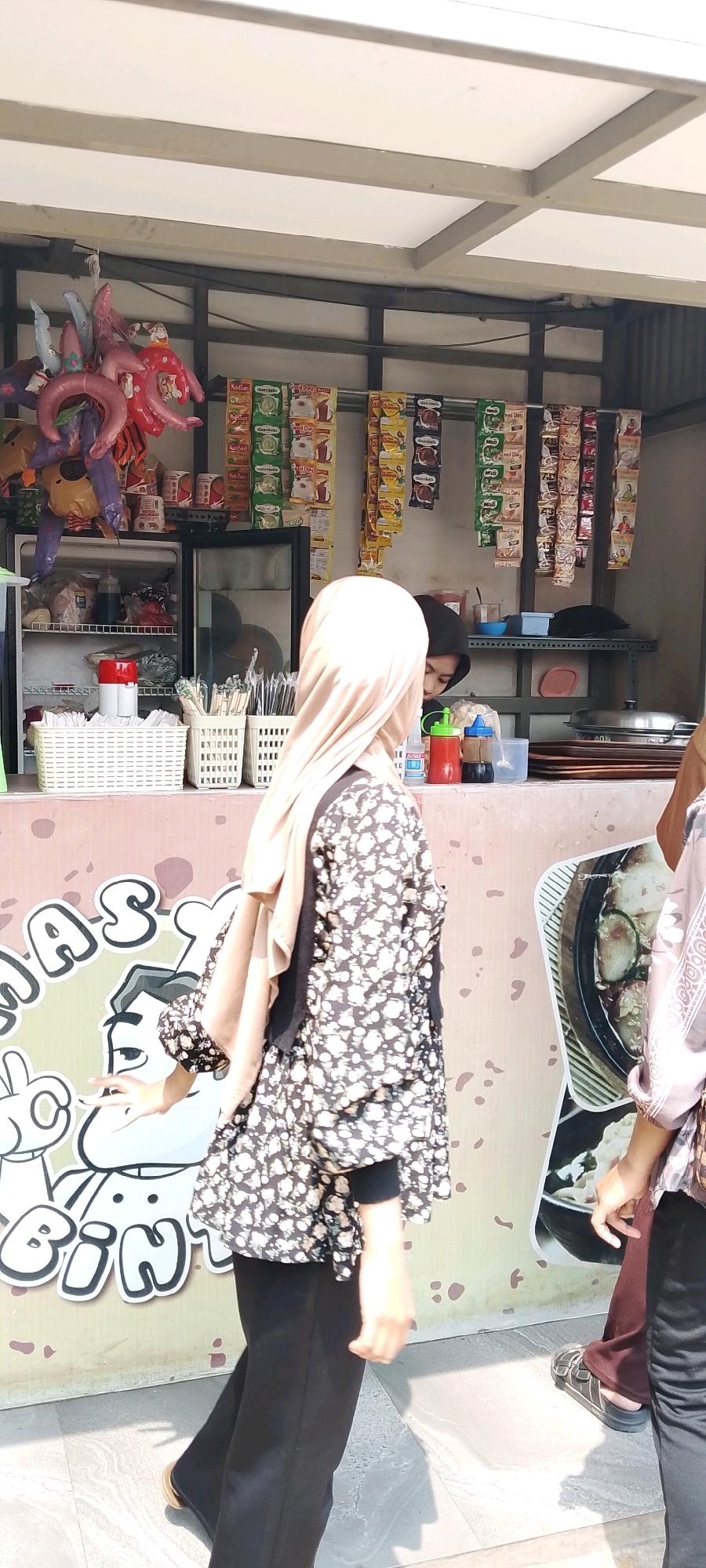 In the city of Sriamur, Indonesia, an outdoor food stall is depicted. A woman wearing a light brown hijab and a black floral print blouse with white flowers, paired with black pants, stands in the foreground, facing right. Behind her, another individual wearing a dark hijab and a dark top is visible, possibly an employee, serving customers. The stall is stocked with various items, including drinks in plastic bottles, snacks in colorful packaging hanging from racks, and a refrigerator containing food items. Baskets filled with disposable cups and cutlery are neatly arranged on the counter. A large graphic illustration is visible on the wall behind the counter, featuring cartoon characters and text that is partially obscured. The ground in front of the stall is paved with light-colored tiles. The overall scene suggests a bustling street food environment during daylight hours.