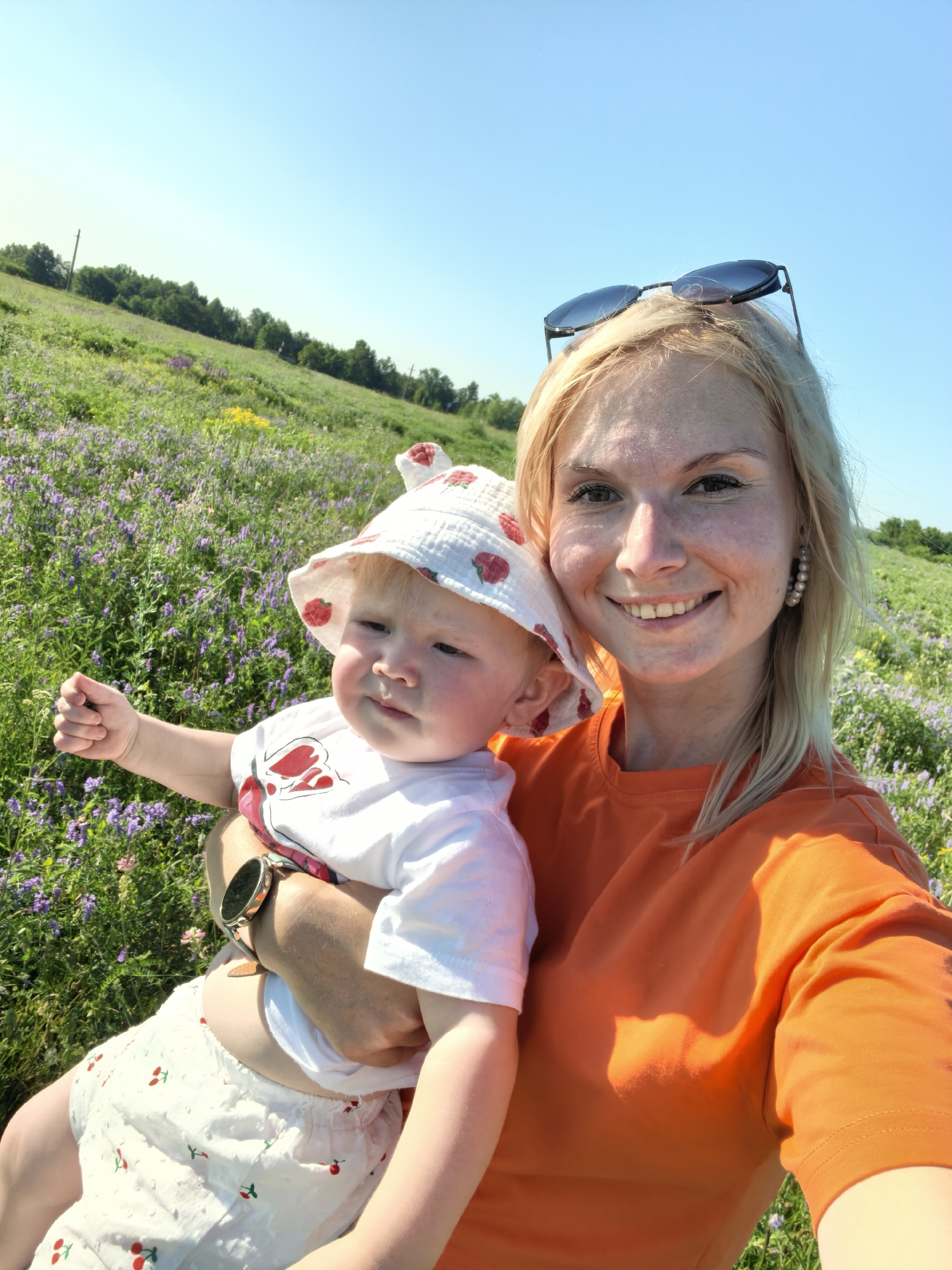 A woman and a child are in a field of purple flowers under a clear blue sky. The woman, on the right, has blonde hair and is wearing an orange t-shirt and sunglasses on her head. She is smiling at the camera. She is holding a child, on the left, who is wearing a white hat with red strawberries and a white t-shirt with a red design. The child is looking to the left and has their hand raised. The field is covered in purple and yellow flowers, with trees in the background. The location is a field outside of Karaiganovo, Russia.
