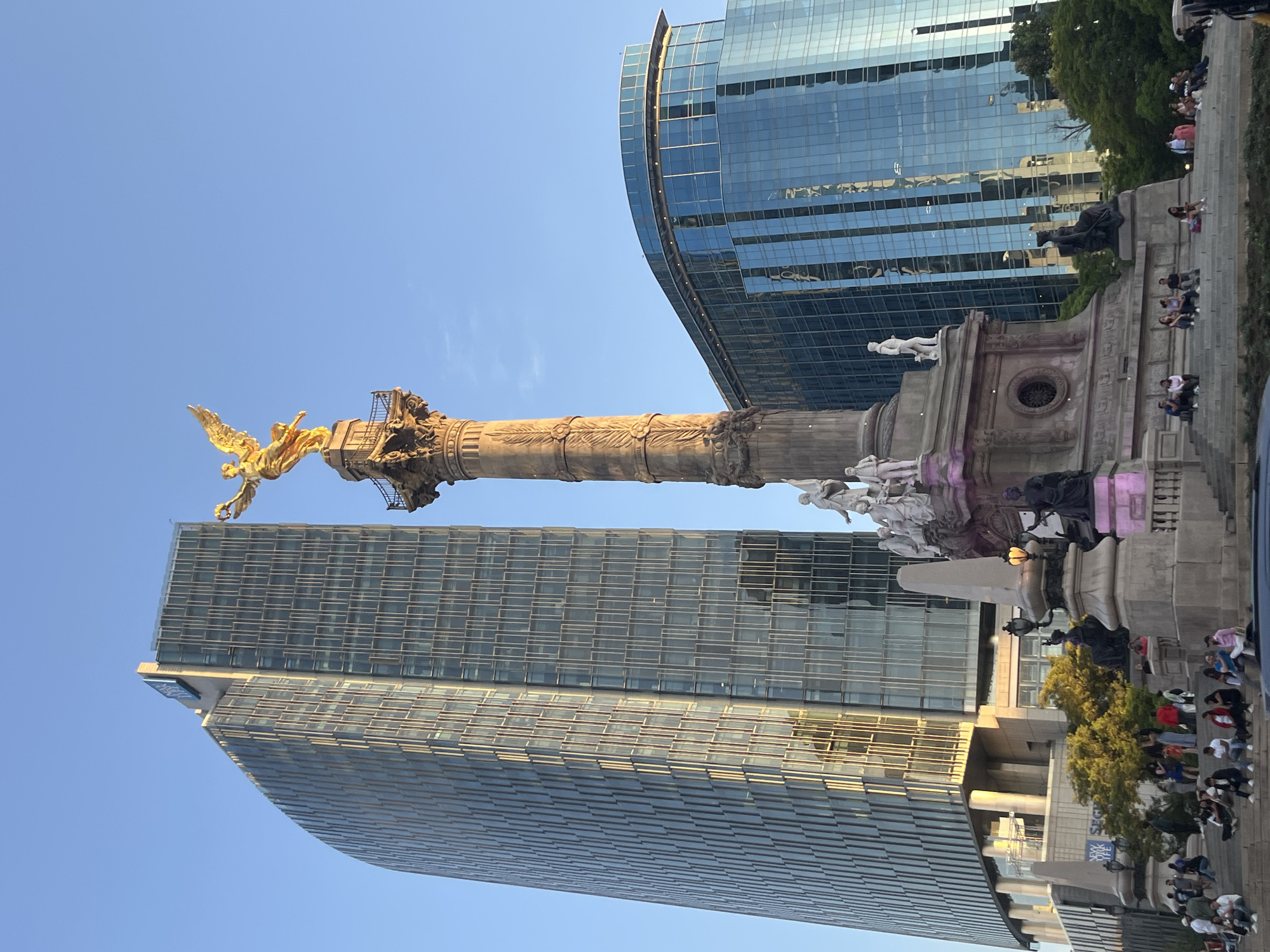 The image shows the Angel of Independence monument in Mexico City, Mexico. The monument is a tall column topped with a golden statue of Winged Victory. The base of the monument has sculptures and is surrounded by steps where people are sitting and standing. Two modern glass buildings are behind the monument, contrasting with the historic architecture. The sky is clear and blue.