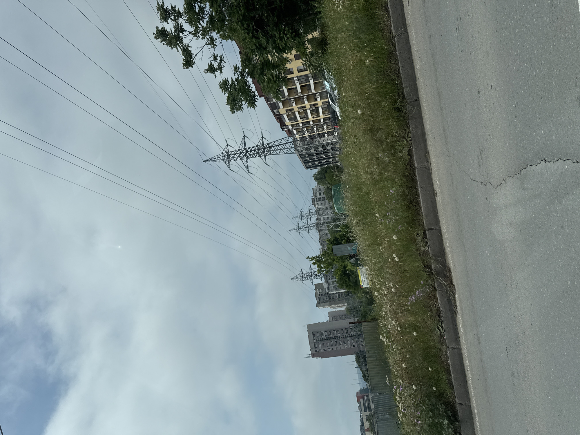 The image presents an urban or suburban scene under an overcast sky. A gray asphalt road occupies the right side of the frame, featuring a concrete curb that separates it from a wide grassy verge. This grassy area is densely covered with weeds and scattered small white and purple flowers, resembling dandelions and other wild blooms.
Dominating the left and central parts of the image are multiple large metal lattice transmission towers (electricity pylons) that support a complex web of power lines stretching across the sky. Several multi-story buildings are visible in the background and mid-ground. One prominent building on the left exhibits a yellow facade with numerous windows, many of which have external air conditioning units attached. Other less distinct gray and white residential or commercial buildings are interspersed among green trees further in the distance.
The sky is bright but uniformly cloudy, suggesting a daytime scene, possibly mid-day or afternoon, without direct sunlight. Subtle lens flares are noticeable in the sky. No individuals or specific activities are depicted, giving the scene a static, objective quality. The asphalt road shows visible cracks, particularly near the curb. While signs are present in the distance within the grassy area, any text on them is illegible. The overall environment combines infrastructure with natural elements and urban development, characteristic of a developed area such as Varna, Bulgaria.