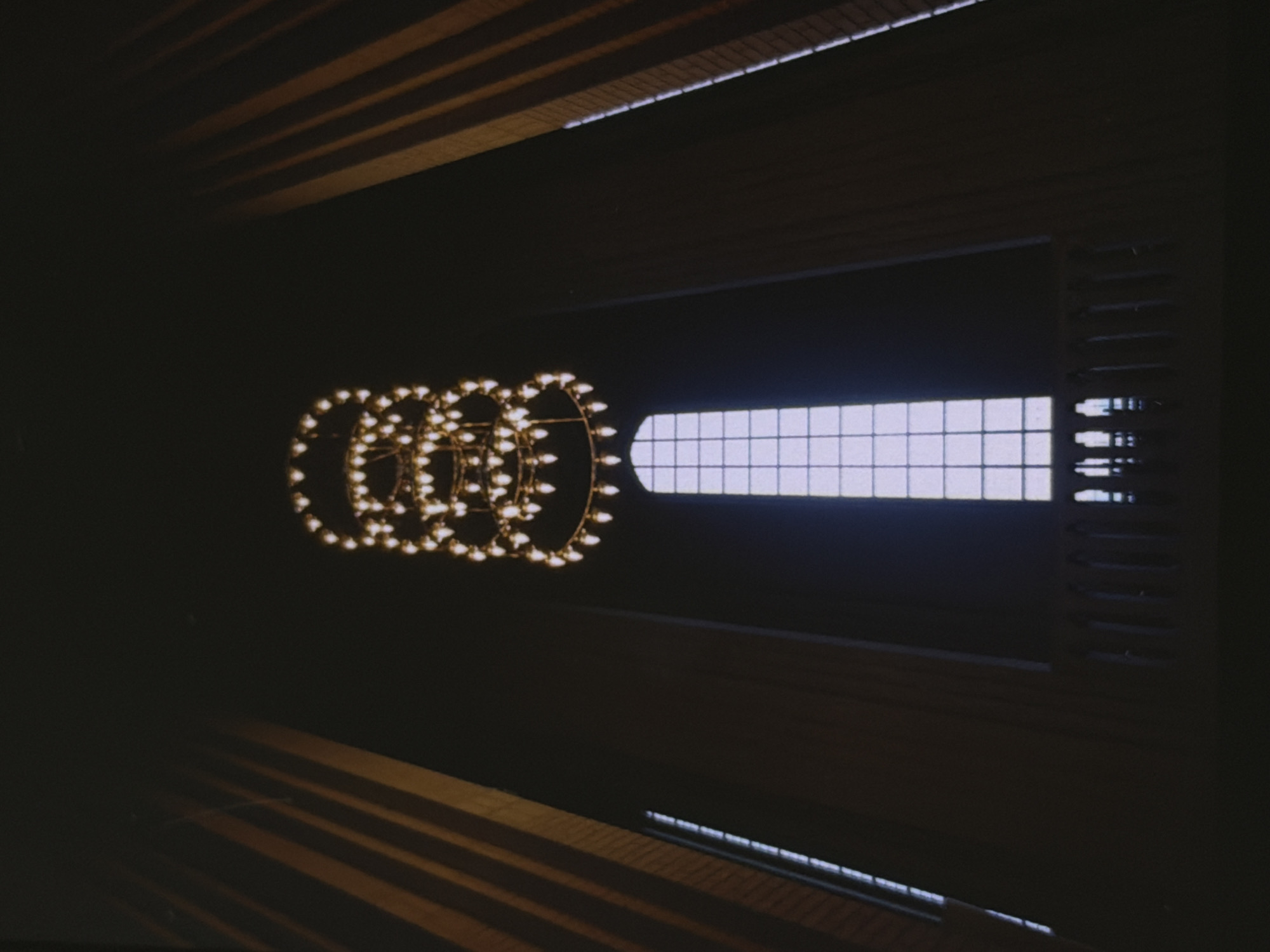 The image shows a low-angle view of a large, ornate chandelier hanging in a dark, cavernous space. The chandelier is multi-tiered and made of a metallic material, possibly brass or bronze, with numerous small, light-colored bulbs illuminating it. Directly below the chandelier is a tall, rectangular window with a grid-like pattern, letting in a soft, diffused light. On the lower part of the image, a decorative railing with vertical supports can be seen, suggesting a balcony or mezzanine. The walls on either side of the window are clad in what appears to be dark wood paneling or drapery, with vertical lines indicating architectural features or folds. The overall atmosphere is one of grandeur and solemnity, with the dim lighting emphasizing the dramatic scale of the architecture and the chandelier. The location is identified as Unknown, Unknown.