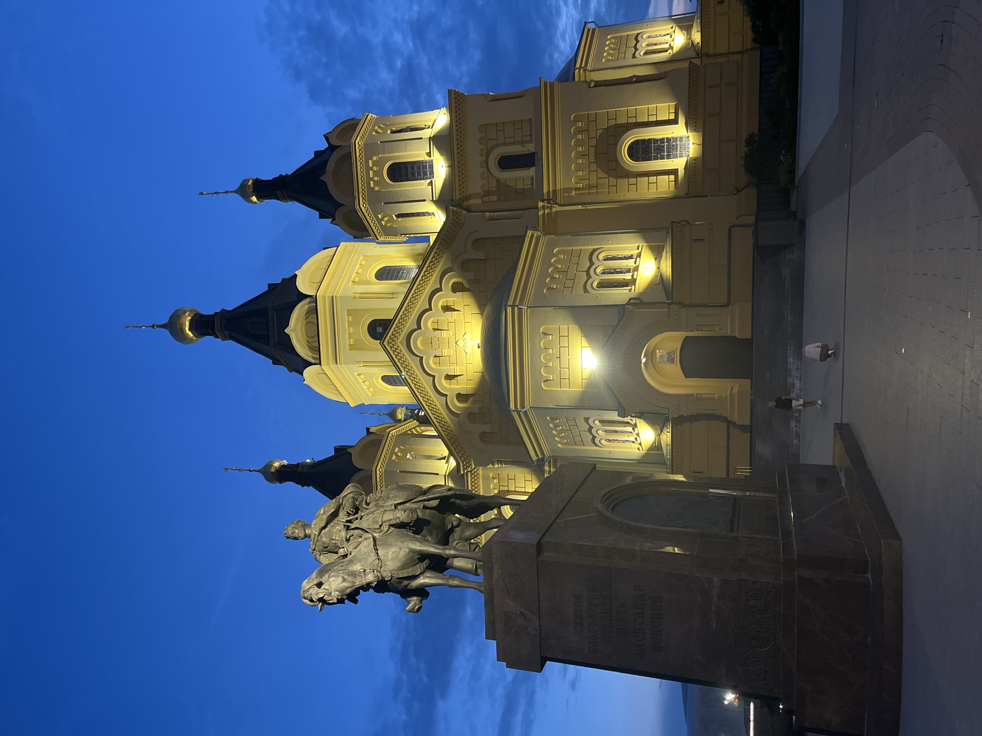 The image depicts the Alexander Nevsky Cathedral in Nizhny Novgorod, Russia, at dusk. The cathedral, illuminated by warm lights, stands prominently with its golden domes and yellow facade against a deep blue sky. In the foreground, a bronze equestrian statue on a granite pedestal is positioned to the left, facing the cathedral. The statue is of Emperor Alexander II. Two individuals, a woman and a child, are visible in the middle ground, near the entrance of the cathedral, with another person further to the right. The ground in front of the cathedral is paved. The overall scene conveys a sense of historical grandeur and civic space.