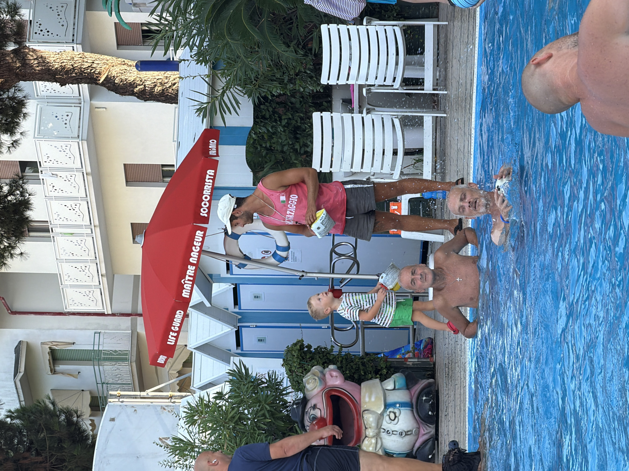 A group of people are in and around a swimming pool at a beach resort in Gatteo, Italy. The pool is bright blue and rippling. In the background, there are white beach huts with blue trim, a red umbrella displaying "LIFE GUARD" in multiple languages, white lounge chairs, and lush green foliage. A large, colorful clown statue is visible near the edge of the pool.
Several men are in the pool; one is looking at the camera and another is holding a white object, possibly a toy, and looking to the side. A young boy in a striped shirt and green shorts stands by the pool's edge, looking up at a man. The man, wearing a pink tank top and grey shorts, appears to be a lifeguard, as indicated by the text on his shirt and the umbrella. He holds a white box in his hands. Another man, wearing a dark t-shirt and shorts, stands to the left of the scene, facing away from the camera.
