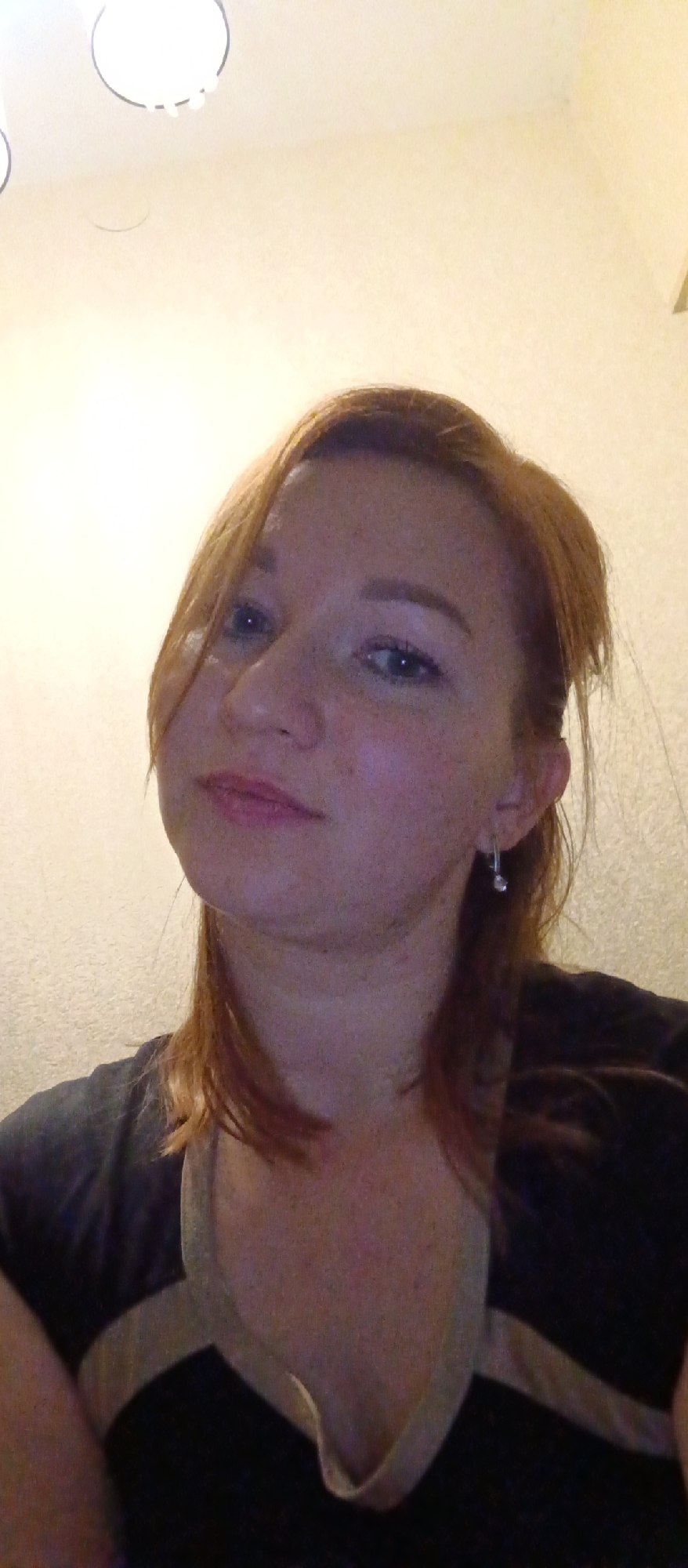 A selfie-style, close-up shot depicts a fair-skinned woman with shoulder-length reddish-brown hair. She has light eyes and is wearing an earring on her left ear. She is wearing a dark t-shirt with a lighter-colored trim around the neckline. The background is a light-colored wall with a lighting fixture visible in the upper portion. The location in the city of Unknown, Unknown cannot be determined from the visual data.