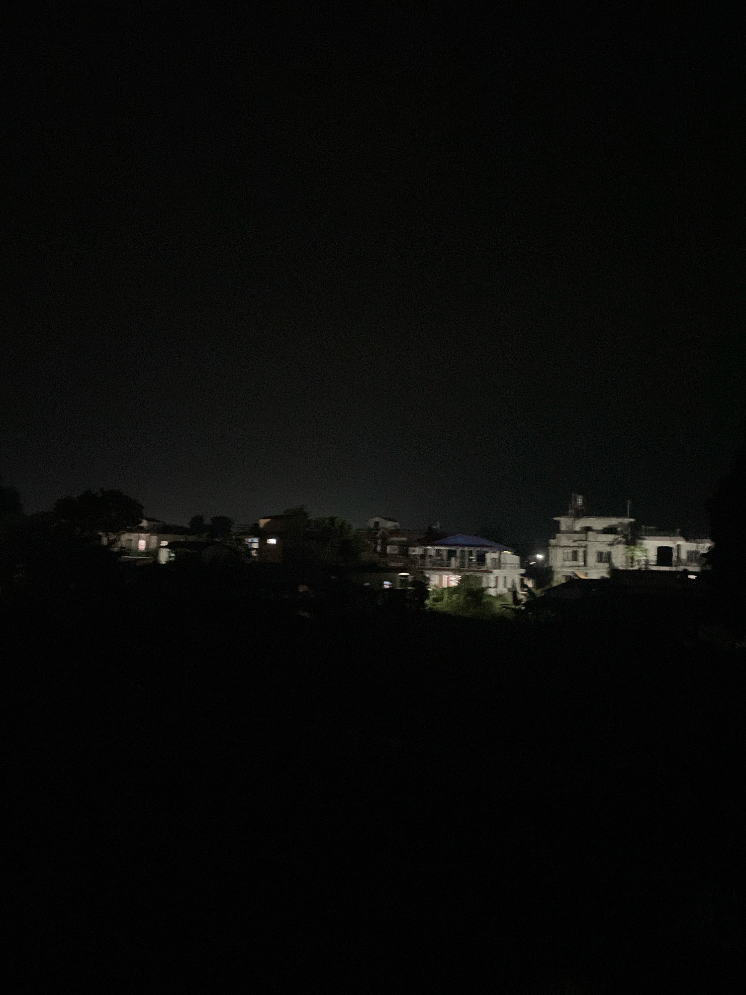 The image captures a nighttime view of buildings in Ratnanagar, Nepal. The scene is dominated by darkness, with artificial lights illuminating several structures. On the left, a building with lit windows is visible amongst trees. To the right, a larger, multi-story building with lit windows and a light on its rooftop stands out. The overall impression is of a residential or commercial area at night, with distinct architectural styles of the buildings suggesting a South Asian context.