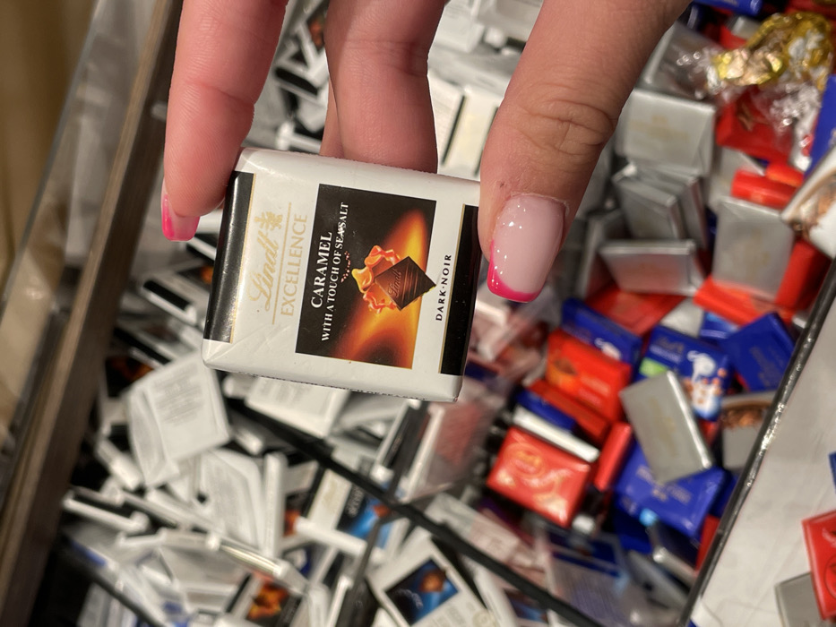 In Marratxí, Spain, a close-up image depicts a human hand holding a single, individually wrapped Lindt Excellence "Caramel with a Touch of Sea Salt Dark-Noir" chocolate square. The chocolate wrapper is predominantly white, featuring the Lindt logo and product description, with a graphic of caramel flowing over a dark chocolate piece. The hand, likely female, displays a manicured index finger and thumb, both with pink French tip nail polish. The chocolate is held above a large, deep bin or container, which is filled with an assortment of various individually wrapped chocolates. These background chocolates are largely out of focus but reveal a mix of white, red, blue, and silver wrappers, suggesting multiple flavors or brands, with many appearing to be additional Lindt squares.