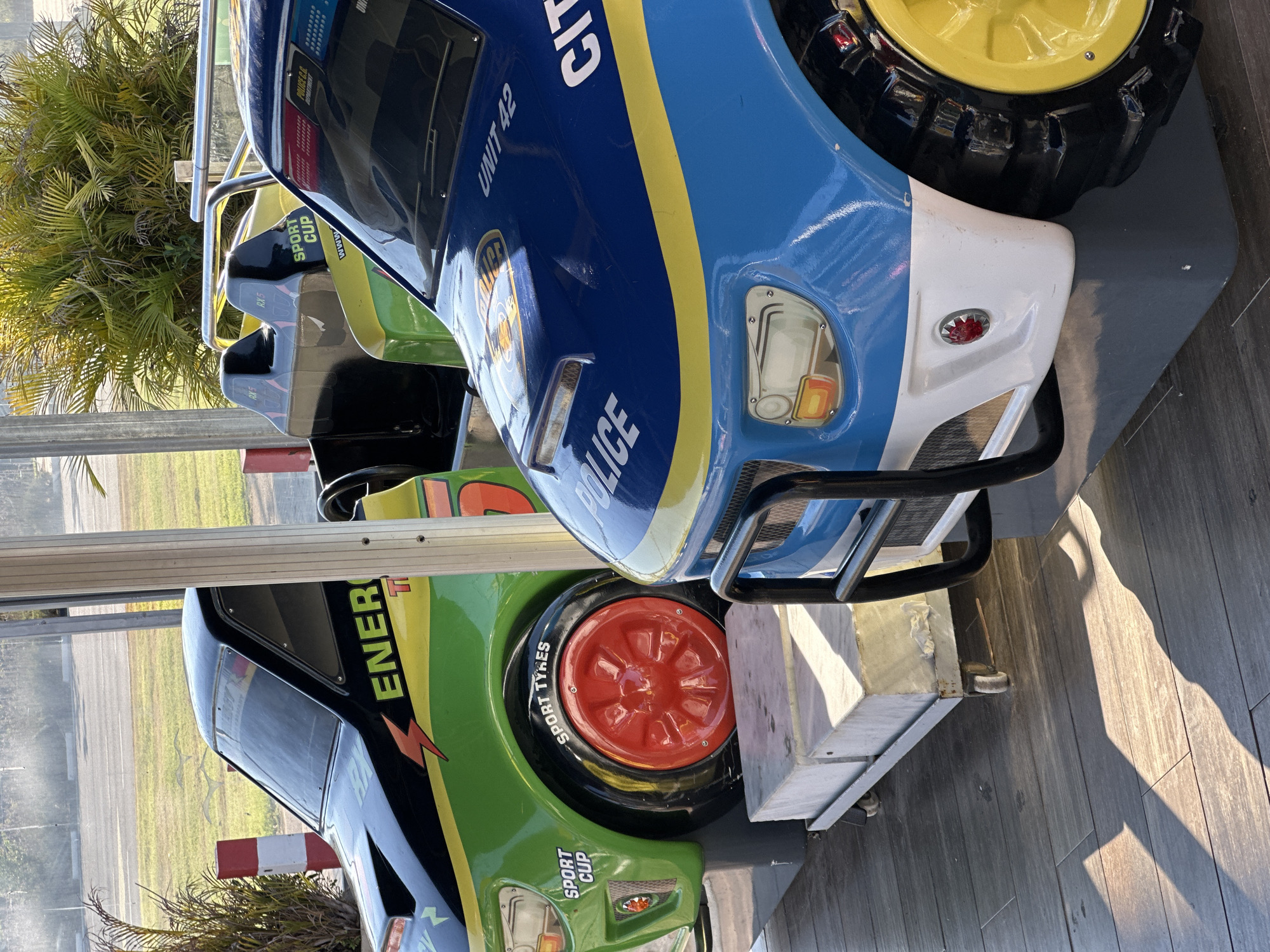 The image shows two colorful kiddie rides, designed to resemble race cars and a police car, parked on a wooden floor outdoors. The ride on the left is green and black with "ENERGY" and a lightning bolt motif. It has a red wheel with "SPORT TYRES" written on the tire. The ride on the right is a blue and yellow police car with "POLICE" and "UNIT 42" visible. The rides are positioned near each other under a covered area, with tropical plants and a paved area in the background. Sunlight casts long shadows across the wooden floor. There are no people visible in the image. The scene appears to be from an amusement area or park. Given the specific location details provided, this scene is located in the city of Hod HaSharon, Israel.