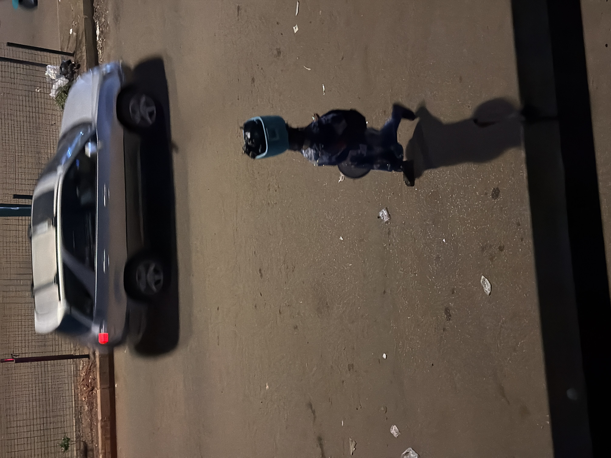 In Bukuru, Nigeria, a person walks along a paved street, carrying a blue container filled with items on their head. The individual is dressed in dark clothing, possibly blue. A dark circular object, perhaps a bag or another container, is held in their left hand. The ground is wet and scattered with debris. In the background, a silver SUV is in motion, its rear brake light illuminated. The scene appears to be outdoors, possibly at dusk or in the evening, with a fence and a wall forming the backdrop to the street.