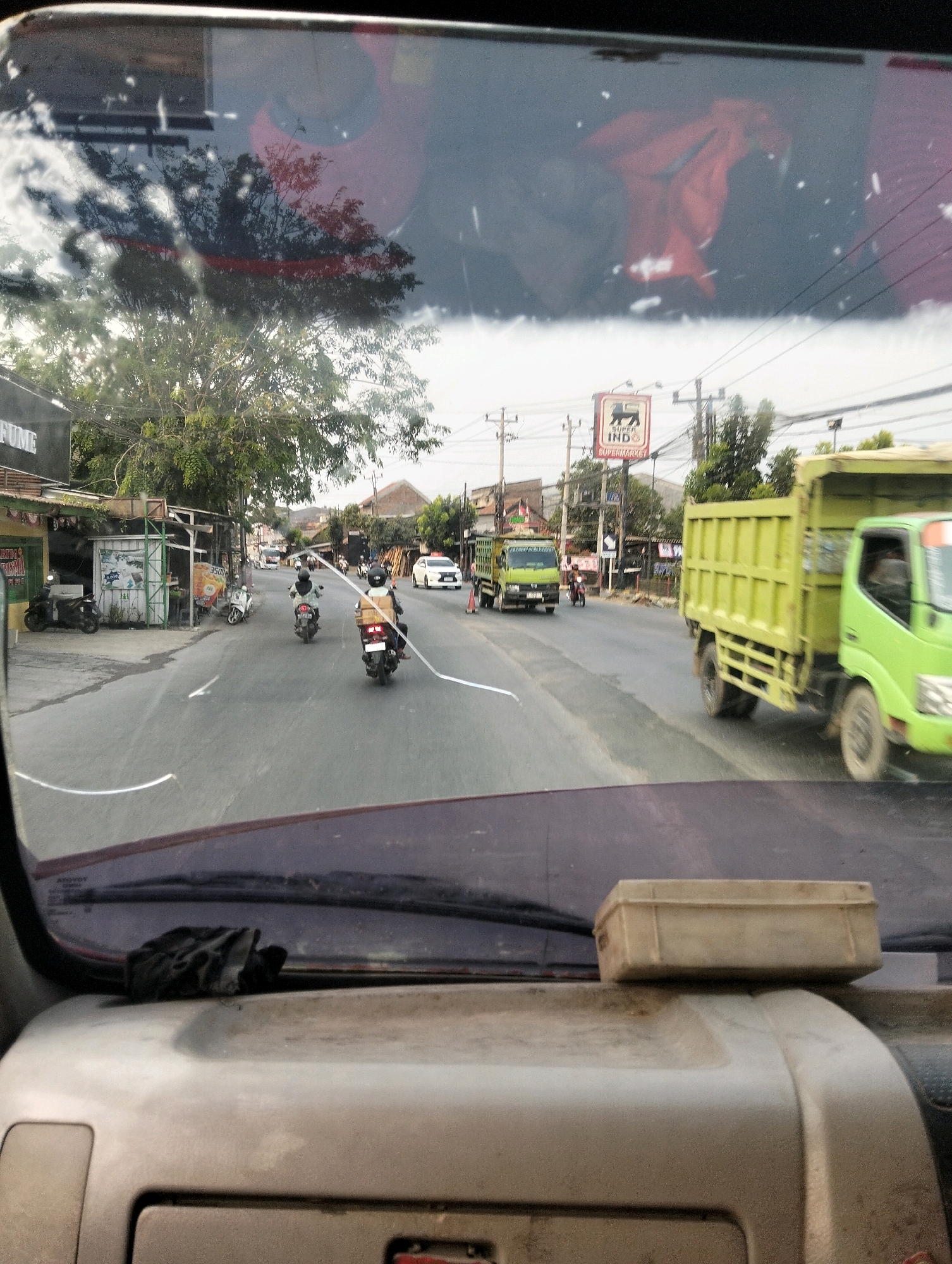 The image is a view from the interior of a vehicle, looking out through the windshield. It shows a street scene in Semarang, Indonesia. In the foreground, on the dashboard, is a black glove and a small, rectangular, light-colored box.
The street appears to be a two-lane road with moderate traffic. Several motorcycles are visible, with riders wearing helmets. A white car is also present on the road. On the right side of the road, a large, bright green dump truck is driving towards the viewer. Further down the road, another green dump truck is visible.
On the left side of the street, there are buildings with storefronts. One sign reads "PERFUME". There are also trees and utility poles. A sign for "SUPER INDO SUPERMARKET" is visible in the distance. The sky is overcast. The overall impression is a typical urban street scene in a tropical climate.