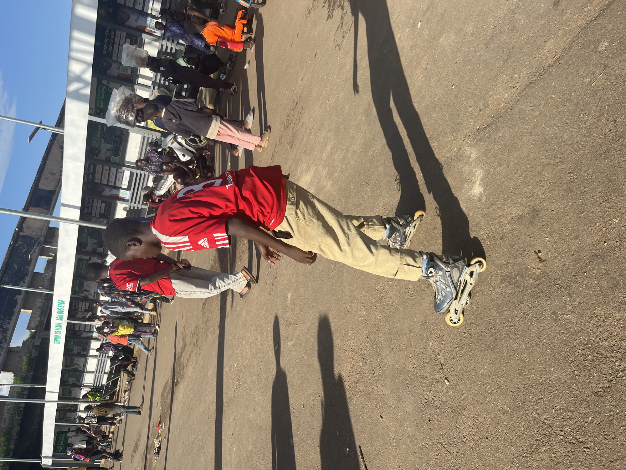 A young boy in a red shirt and tan pants is rollerblading on a dirt surface in front of a bus stop shelter labeled "TERMINUS MAIN ARK BUS STOP." He is facing away from the camera, and the sun is casting a long shadow to his left. Behind him, several other people are milling about, some sitting and others standing. Some individuals are wearing colorful clothing, and there is a motorcycle parked near the bus stop. The environment appears to be an outdoor, urban setting, possibly a marketplace or transportation hub. The bus stop shelter has signage with information and graphics.