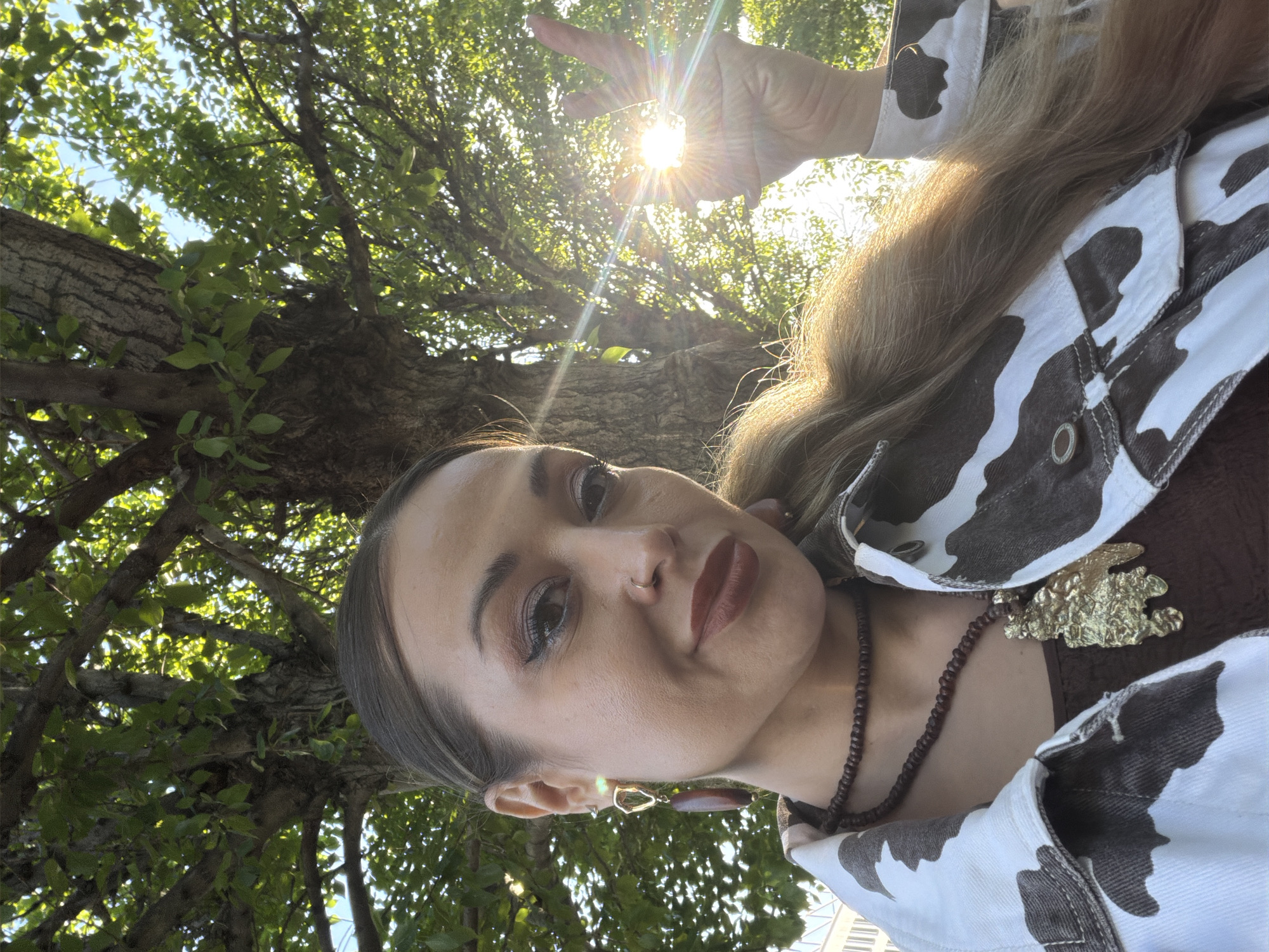 A person, presumably female based on facial features and jewelry, is taking a selfie while standing under a tree. The sun is visible through the tree branches and the person's hand, which is raised in a peace sign. The person is wearing a cow-print jacket and several pieces of jewelry including a necklace and earrings. The location appears to be outdoors, under a tree. There is no discernible information to indicate the exact location or city. The suggestion it is in Krasnoyarsk, Russia is not confirmed by the image.