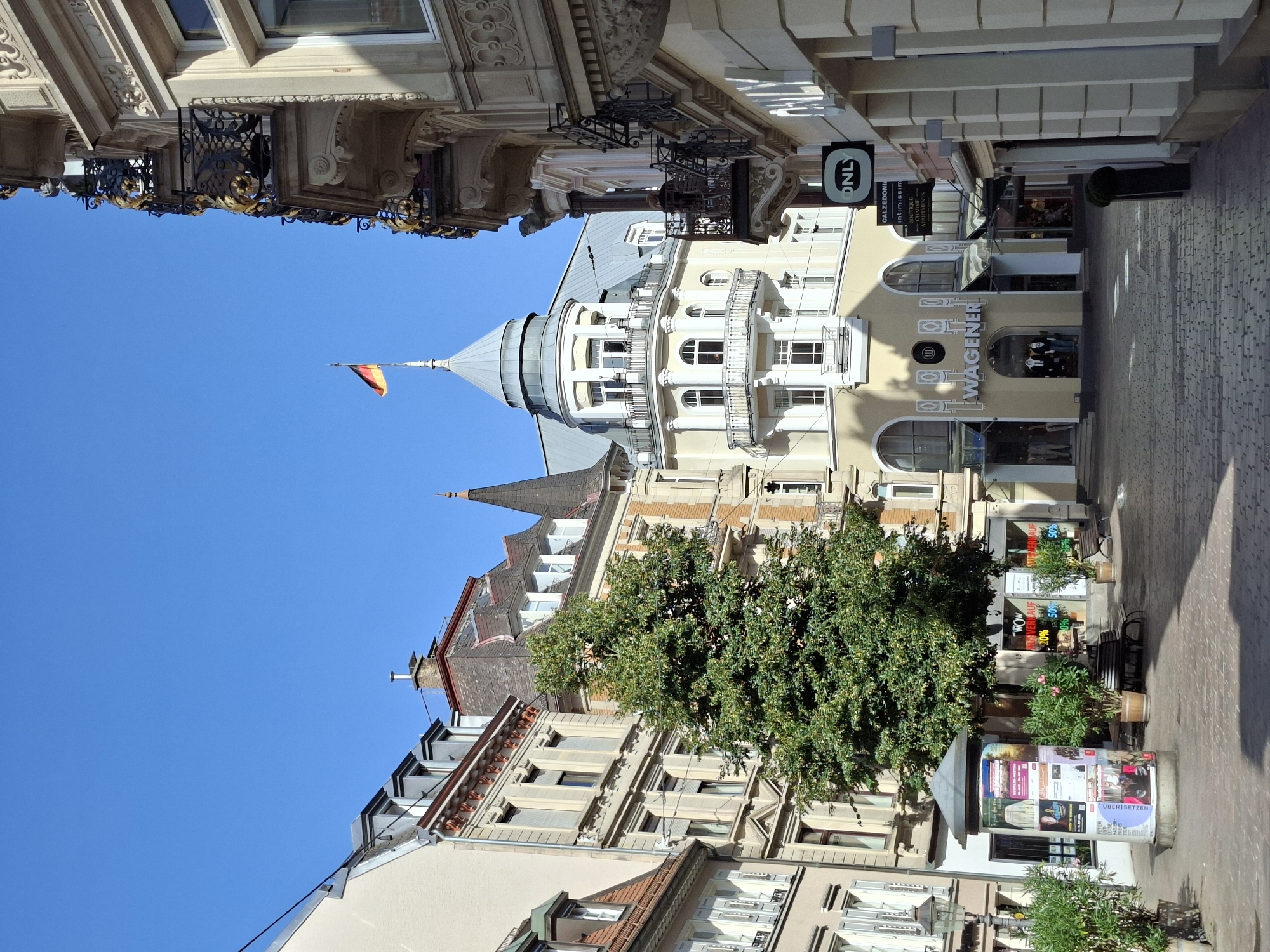 In Baden-Baden, Germany, on a bright, sunny day, a cobbled pedestrian street is lined with ornate European architecture. In the center of the frame, a large, mature tree provides shade. On the left, a cylindrical advertising pillar displays various posters. Further down the street, businesses with storefronts such as "WAGENER," "ONLY," "CALZEDONIA," and "INTIMISSIMI" are visible. The buildings exhibit classical architectural styles with balconies, intricate stonework, and large windows. A German flag flies atop a tower. A few people are seen on balconies and in the street, with benches scattered throughout the area. The sky is clear and blue.