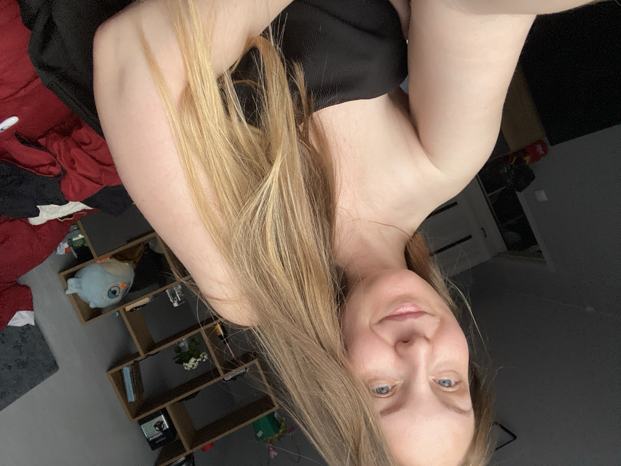 The image is a self-portrait featuring a woman with long, light blonde or light brown hair, which cascades over her shoulder and arm. She has light-colored eyes and a gentle, soft smile, looking directly at the camera. She is wearing a dark, possibly black, strapless top and a delicate gold-colored chain necklace is visible around her neck.
The setting is an indoor room, likely a personal space such as a bedroom or living area in Noviy, Russia. In the background to her left, crumpled red and dark fabrics are visible, suggesting bedding or clothing. To her right, a dark wall and a door frame are present. Behind her head and shoulder, a modular wooden shelf unit with multiple compartments holds various items. These include a light blue, spherical plush toy resembling an owl or bird, books, and small decorative objects, including what appears to be a white flowering plant. The lighting in the room is soft and ambient, without harsh shadows, suggesting general indoor illumination. There is no visible text in the image.
