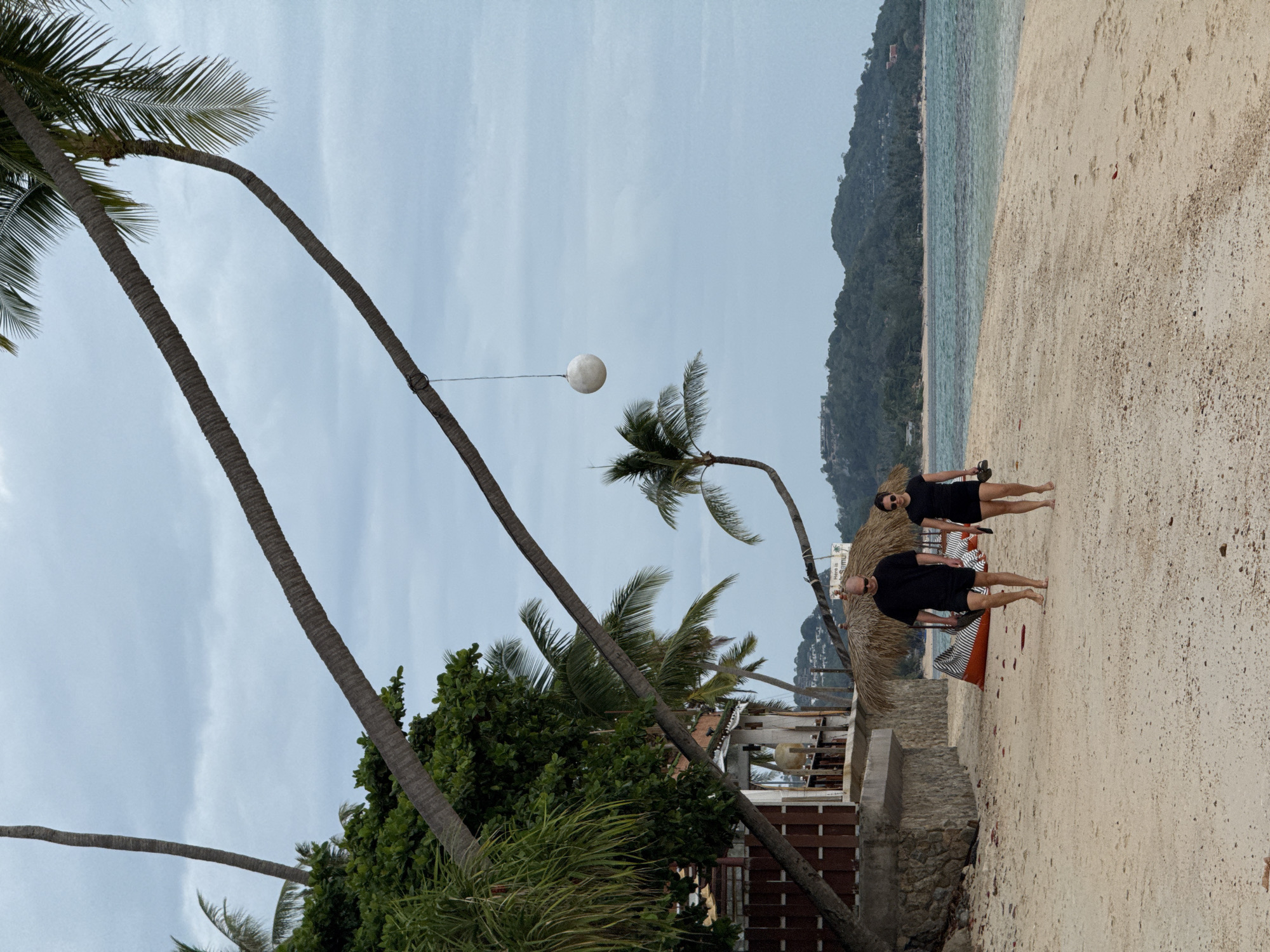 On a cloudy day at a beach in Ko Samui District, Thailand, a bald man and a woman with dark hair are walking barefoot on the sand towards the right of the frame. Both are wearing black t-shirts and black shorts. The man is carrying black flip-flops in his right hand, and the woman is carrying black flip-flops in her left hand. Behind them, there are two thatched-roof umbrellas with striped cushions underneath. To the left, a building with a stone base and a wooden facade is partially visible, with palm trees and lush greenery surrounding it. Several palm trees are prominent in the foreground and midground, with their fronds reaching towards the overcast sky. A white spherical object hangs from a rope tied to a palm tree trunk. In the background, a calm turquoise ocean meets a densely forested hill.