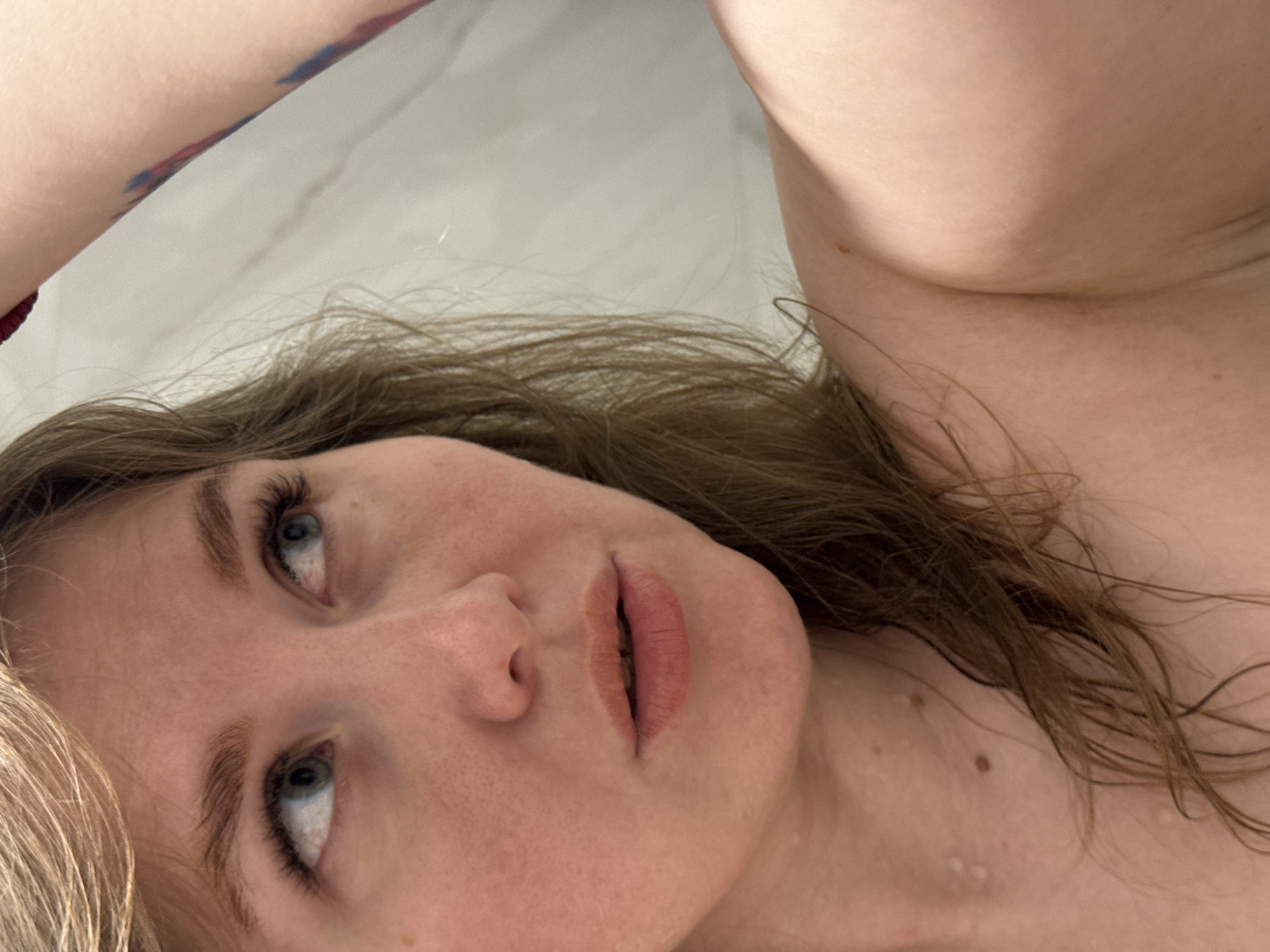 A close-up, tilted, and slightly out-of-focus shot reveals a woman's face and partial upper body, with light-colored skin. She has light brown hair, blue eyes, and pink lips. Water droplets are visible on her face and chest, suggesting she may have recently bathed or showered. Part of an arm with a tattoo is visible in the upper left corner of the frame. No elements indicate the city of Ферма, Russia.