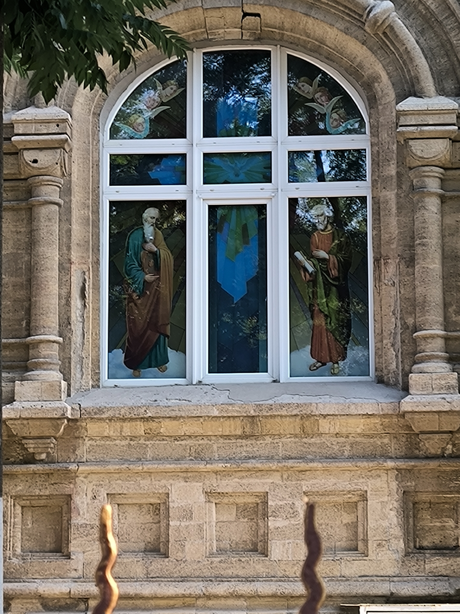 The image shows a stained-glass window with religious figures, set within a stone building. The window is arched at the top and divided into several panes by white mullions. The upper arched section of the window depicts three angels. Below the angels, there are three vertical panes. The left pane shows a male figure with a beard, wearing a long robe and holding an object. The central pane has abstract blue and green stained glass. The right pane depicts another male figure with a beard, wearing a robe and holding a scroll. The building appears to be made of stone blocks. In the foreground, a portion of a sculpted object with a spiral design is visible. The environment appears to be outdoors, with some greenery visible at the top left of the image. The image is taken from a frontal perspective, slightly looking up at the window.
The specific location is the window of the Roman Catholic Church of the Assumption of the Blessed Virgin Mary in Mykolaiv, Ukraine.