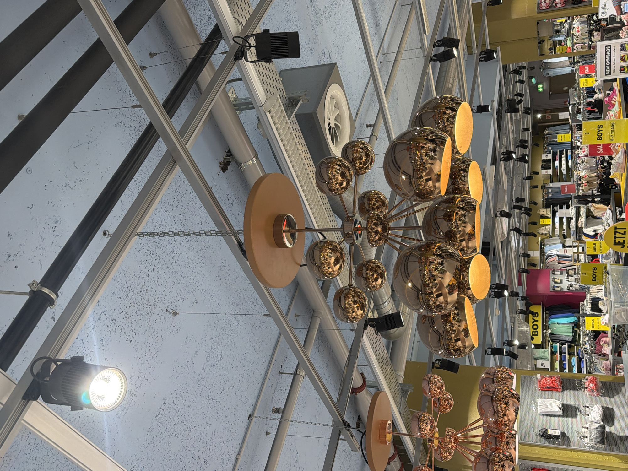 The image depicts a view of a store interior, likely in Spreitenbach, Switzerland, given the German signage. The perspective is looking upwards towards a ceiling with a complex network of metal beams, pipes, and fixtures. Two modern chandeliers with multiple reflective, golden spherical bulbs are prominently featured, one near the center and another towards the lower part of the frame. The lighting is bright, illuminating a retail space in the background where clothing displays and promotional signs for "BOYS" and "GIRLS" sections are visible. Clothing items are neatly arranged on shelves and racks. The environment is commercial, suggesting a retail store, potentially a clothing store in a shopping center or mall.