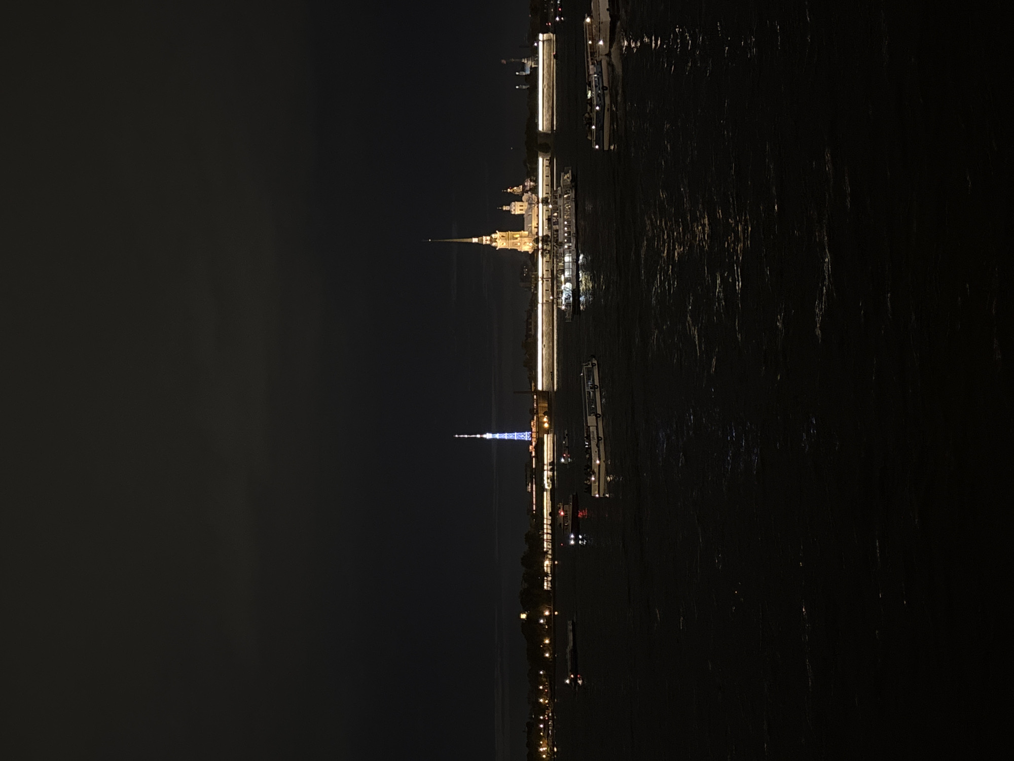 The image depicts a nighttime scene in Saint Petersburg, Russia, featuring illuminated landmarks along a river. The Peter and Paul Fortress is visible, its spire prominently lit in yellow. The image also shows the St. Isaac's Cathedral lit up in blue. The reflection of these lights shimmers on the dark water. Various boats with illuminated decks can be seen along the river. The background is dark, likely representing the night sky. The orientation of the image is rotated.