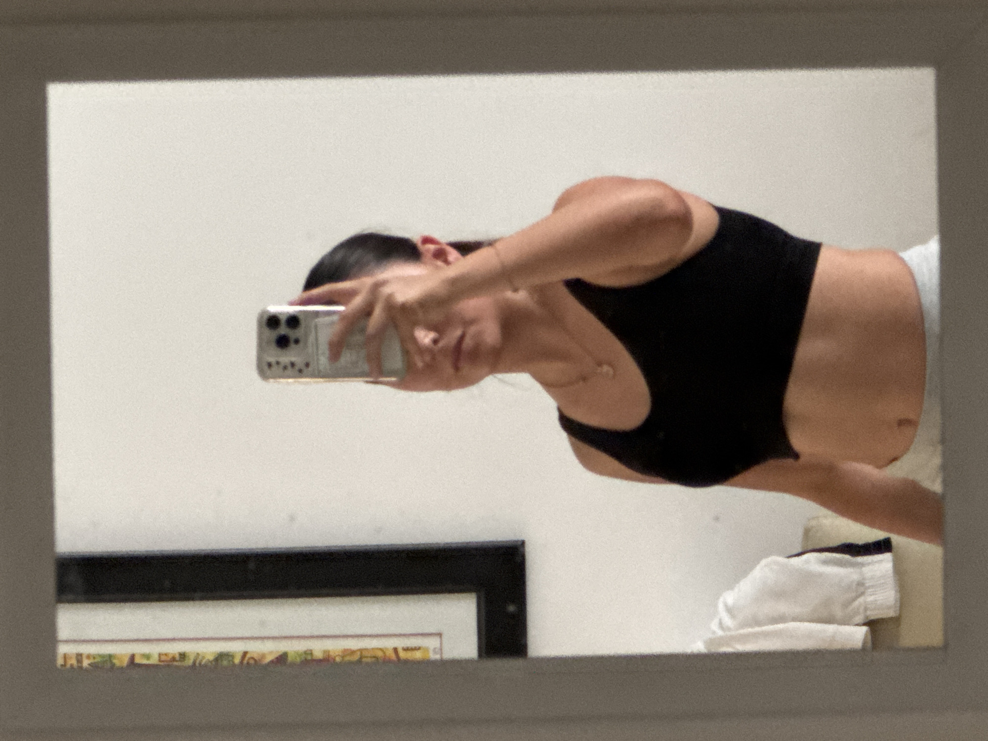 A woman in a black top takes a selfie in a mirror. The background includes a white wall, a framed picture, and a stack of towels. The reflection suggests an indoor environment. The location is not identifiable as San Francisco Ocotlán (Ocotlán), Mexico, from the visual information.