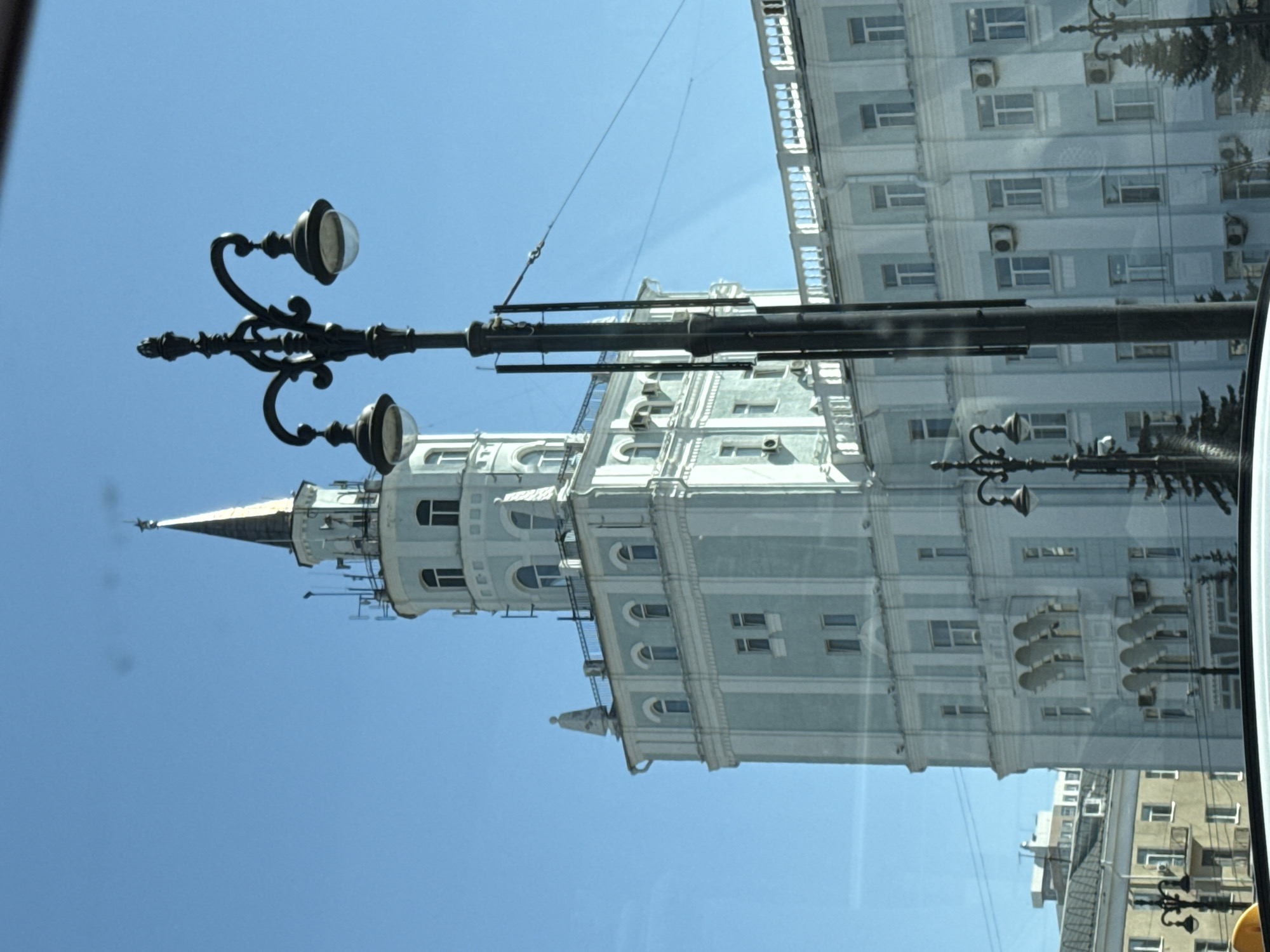 The image, taken from a vehicle in motion, showcases a tall, light-colored building with a pointed spire, potentially of Soviet-era architecture, in Perm, Russia. The building features numerous windows, some with visible air conditioning units. In the foreground, a black, ornate lamppost with globe-shaped lights and overhead electrical wires are visible. The blue sky is partially obscured by the building and the lamppost. The view is captured through a window, possibly a car or bus window, with reflections visible on the glass.