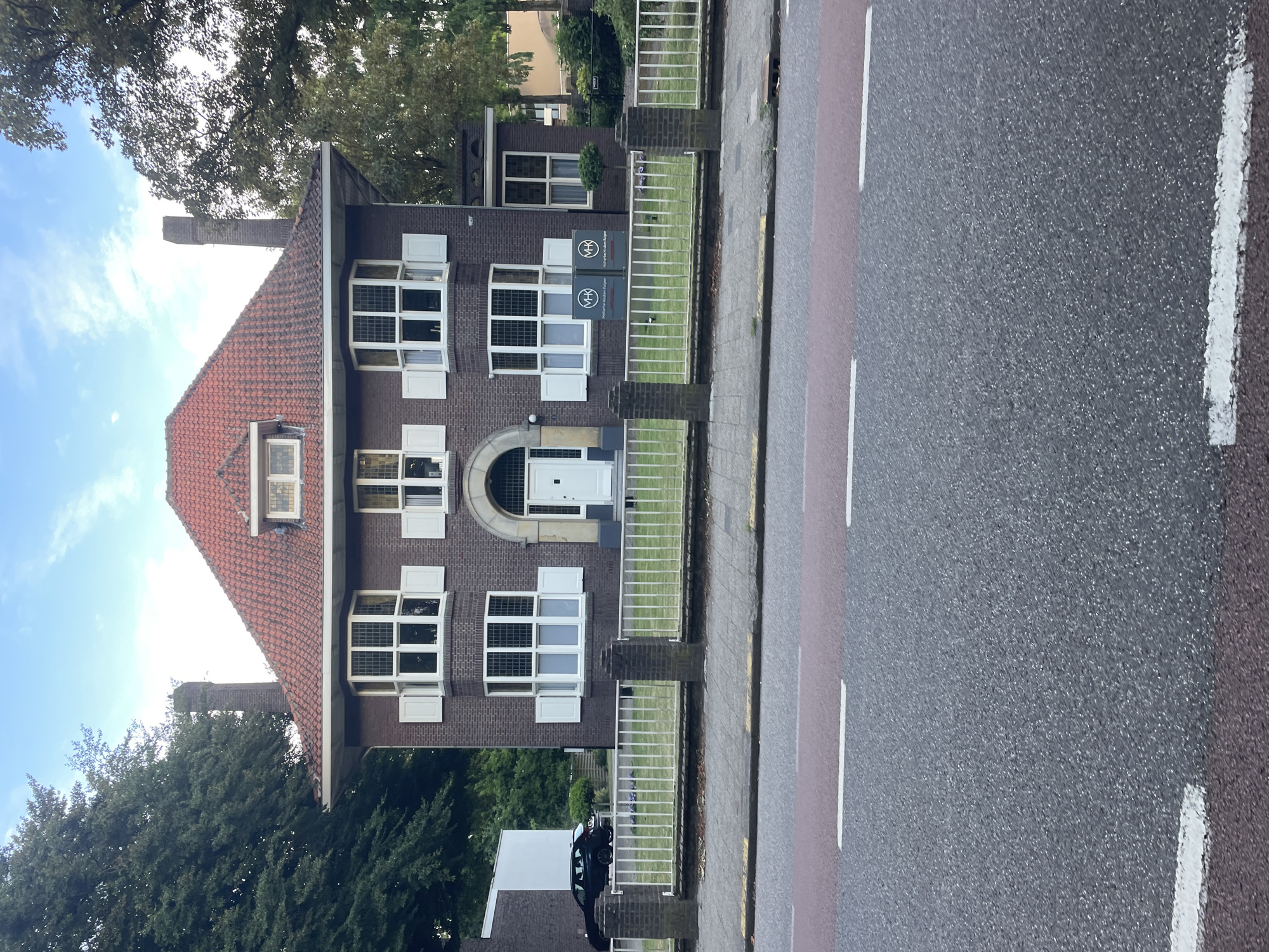 This is a vertical, eye-level, outdoor shot of a brick building with a red-tiled roof. The building appears to be an institutional or office building, with multiple windows, some of which have white shutters. There are two signs on the building with the text "VHK" in a circular logo. The building is next to a sidewalk and a road with white and red lane markings. Trees and a cloudy sky are visible in the background. The scene appears to be set during the daytime, possibly in the late afternoon, with a mild or overcast weather condition. There are no people visible in the image, but a dark-colored car is parked on the property behind the building. The image captures a quiet, orderly scene, suggesting a functional rather than a recreational setting. The location is Weert, Netherlands.