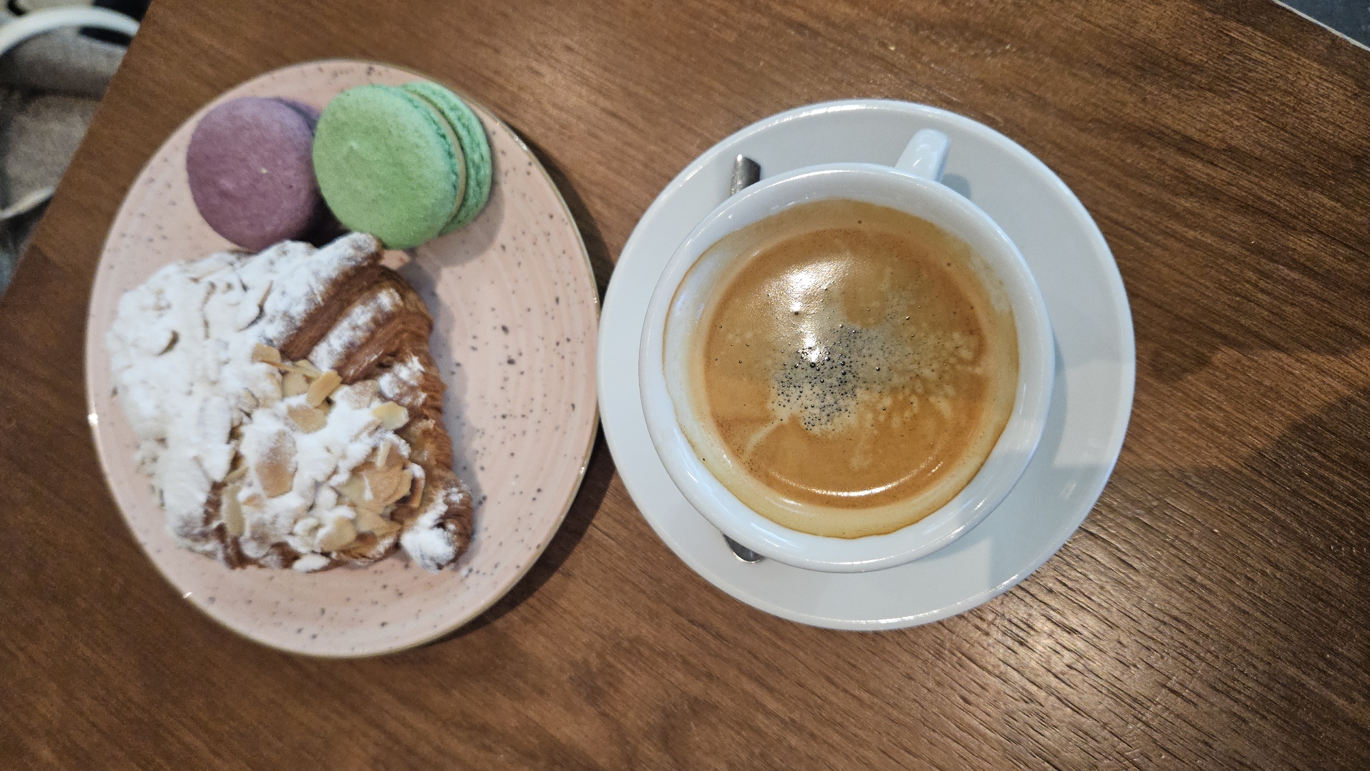 A high-angle, medium shot captures a pastry and two macarons on a pink speckled plate, positioned on a wooden table. To the right of the plate, a white espresso cup filled with dark coffee sits on a matching saucer. The croissant is topped with powdered sugar and slivered almonds. A green and a purple macaron are placed next to the croissant. The wooden table surface displays a visible wood grain pattern. The background is blurred, showing a hint of a chair. The scene is devoid of people, action, or interaction, presenting a static arrangement of food and drink.