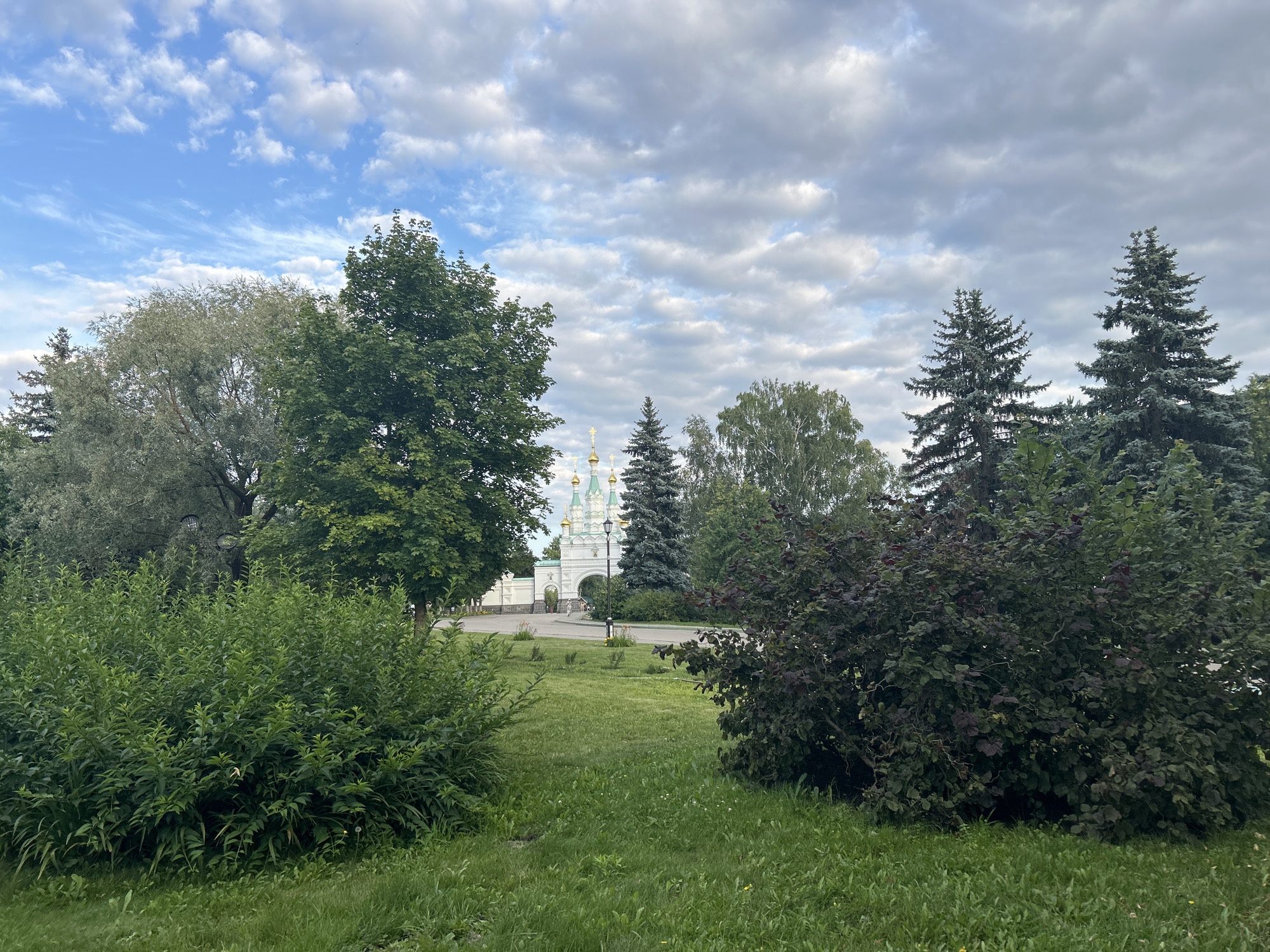 In Diveyevo, Russia, a picturesque scene unfolds with a large white arched gateway leading to a religious complex, characterized by its golden domes. The gateway is flanked by lush greenery, including various trees and bushes. In the foreground, a neatly trimmed lawn area is interspersed with more dense shrubbery. Tall coniferous trees stand prominently in the background, framing the gateway and adding to the natural beauty of the environment. The sky above is a mix of blue and white clouds, indicating a partly cloudy day. A few indistinct figures are visible near the gateway, suggesting a public space. The overall impression is one of serenity and spiritual significance, set within a well-maintained natural landscape.