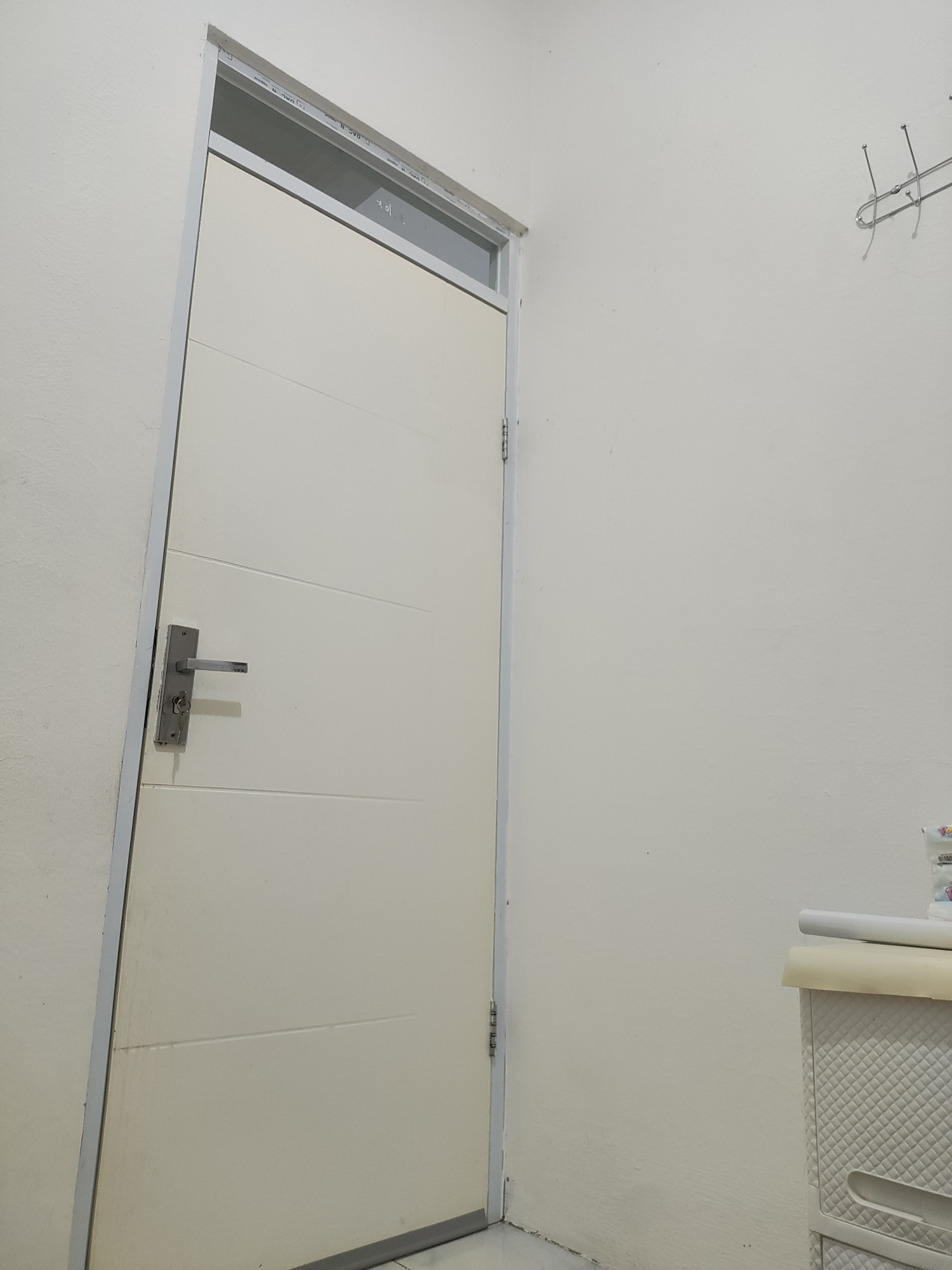 The image depicts an interior scene of a room, likely a hallway or utility space, within a building in Wanajaya, Indonesia. On the left side of the frame is a closed, light-colored door with a silver handle and a key inserted into the lock. The door has horizontal grooves. The door frame is also light-colored and appears to be made of metal. Above the door, there is a narrow horizontal window with a metallic frame, through which a darker interior space is visible. The walls are painted a neutral, off-white color. On the right side of the image, against the wall, is a white, multi-drawer storage unit. On top of this unit are rolls of what appears to be wrapping paper or fabric, one white and one cream-colored. To the upper right of the frame, a metal coat rack with several hooks is mounted on the wall. The lighting is artificial and casts soft shadows.