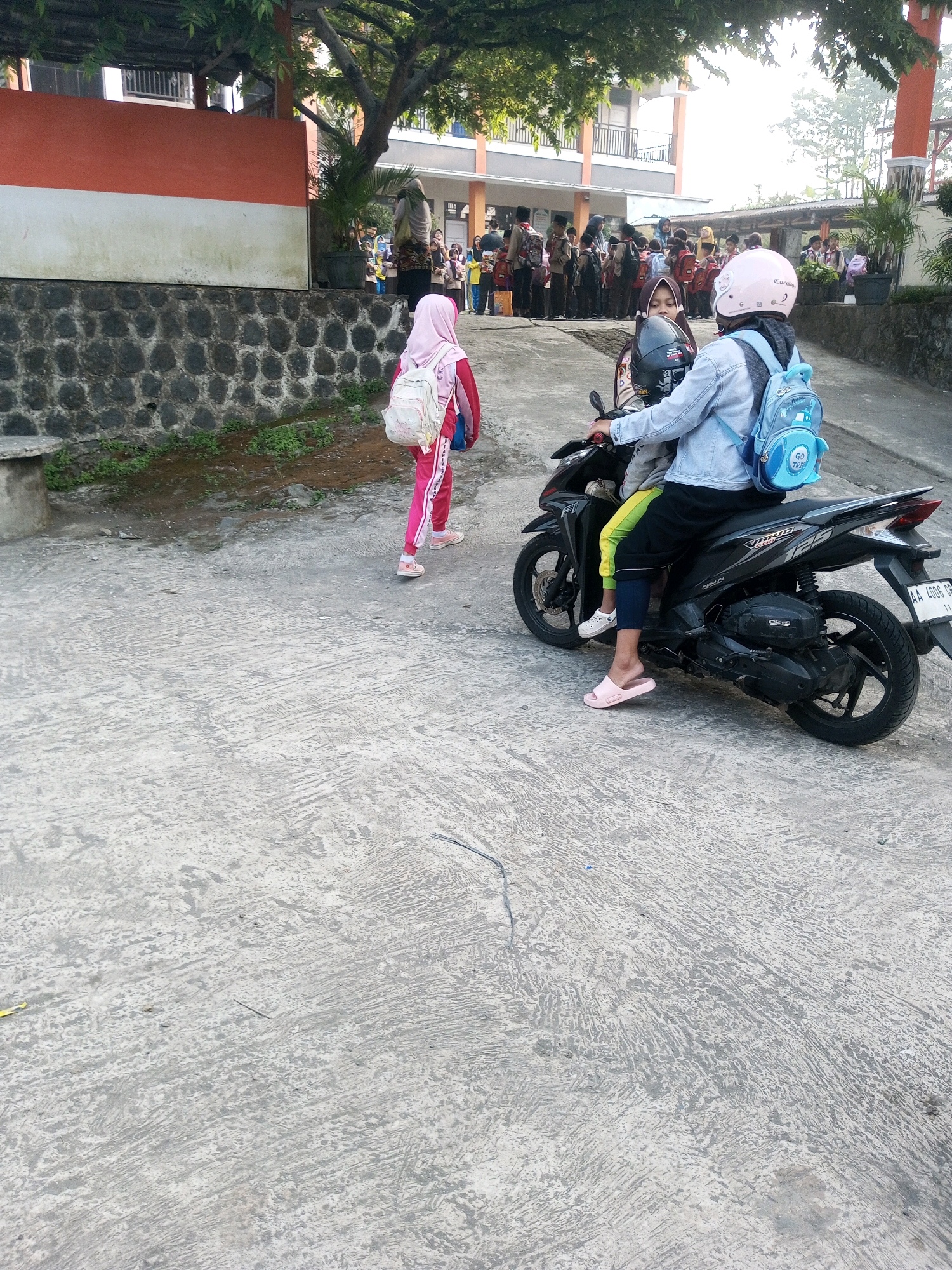 In Krasak, Indonesia, a young girl wearing a pink headscarf and pink track suit, carrying a white backpack, walks away from the camera on a concrete pathway. Behind her, a person on a motorcycle with a pink helmet and a light blue backpack is stopped. The motorcycle rider, wearing a denim jacket, has another person, also wearing a headscarf, seated behind them. Both individuals on the motorcycle appear to be young. In the background, a group of students wearing uniforms and carrying backpacks are gathered near a building with an orange and white facade, surrounded by trees and potted plants. The ground is wet and the overall environment suggests a school setting.