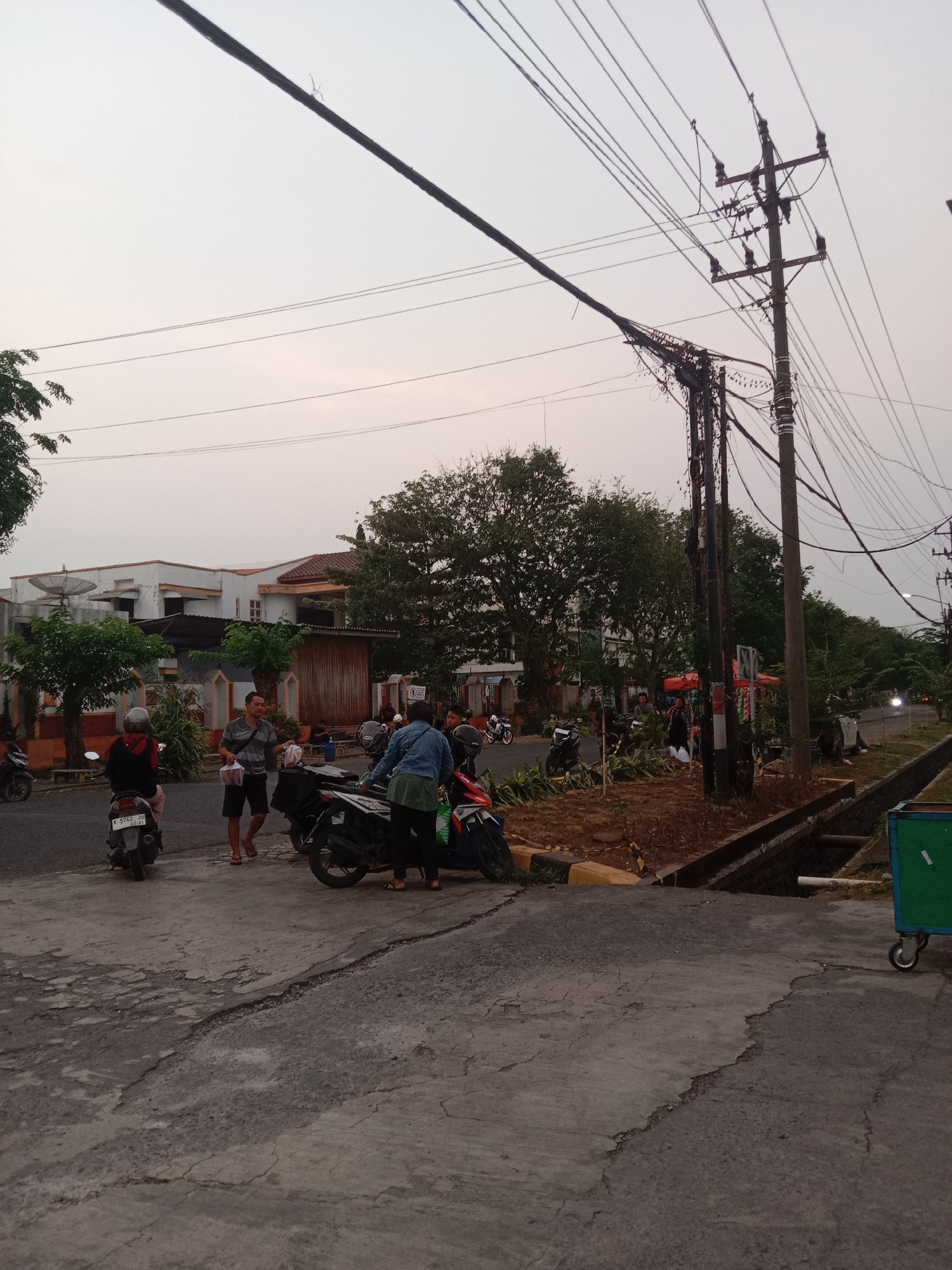 This image depicts an outdoor scene in Semarang, Indonesia, characterized by a street with several people and motorcycles. In the foreground, a cracked asphalt road is visible, with a drainage ditch running alongside it on the right. Further into the scene, a group of individuals are gathered around motorcycles. One man, wearing a grey and black striped shirt and black shorts, holds plastic bags containing what appears to be food. Another person, wearing a blue denim jacket and black pants, stands near a motorcycle. Several motorcycles are parked on the roadside. In the background, a white building with a red-tiled roof is visible, along with trees and utility poles carrying numerous electrical wires. The sky is overcast, suggesting it might be dawn, dusk, or a cloudy day. The overall impression is a casual street scene, possibly a stop for food or rest during a journey.