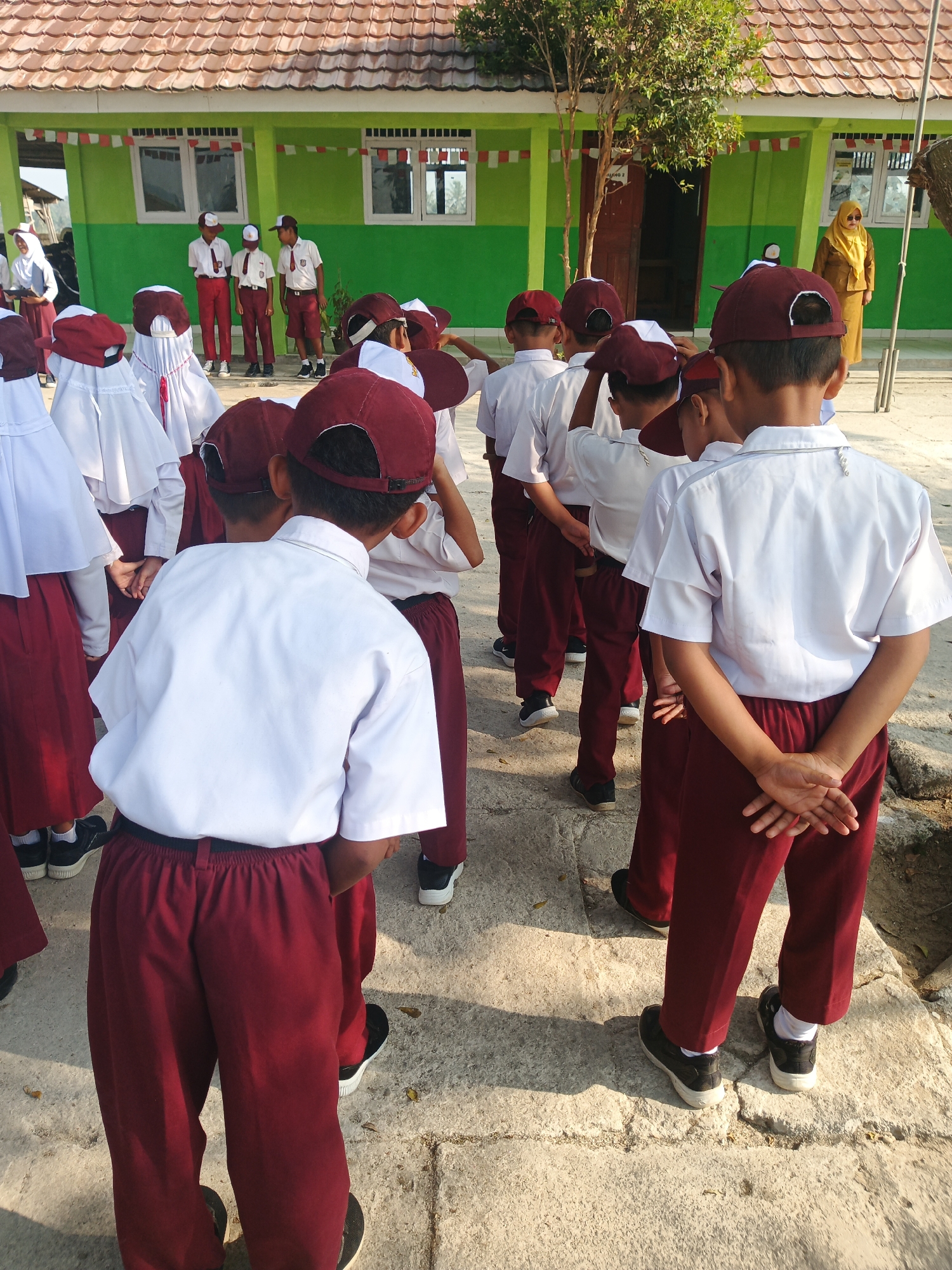 The image shows a group of Indonesian elementary school children, dressed in their school uniforms, gathered outside a school building. The children are wearing white shirts, maroon pants, and maroon baseball caps. Some of the children are facing away from the camera, while others are partially visible from the side or front. They appear to be standing in formation, possibly for a flag ceremony or assembly. The school building is painted green with a brown tiled roof, and there are windows and a door visible. Decorations, including red and white banners, are strung along the facade of the building. A woman in a yellow headscarf and dress can be seen standing near the door of the school. The ground is paved with concrete blocks, and there are some dry leaves scattered around. The sun is shining, casting shadows on the ground. The scene is set in the city of Rejo Basuki, Indonesia.
