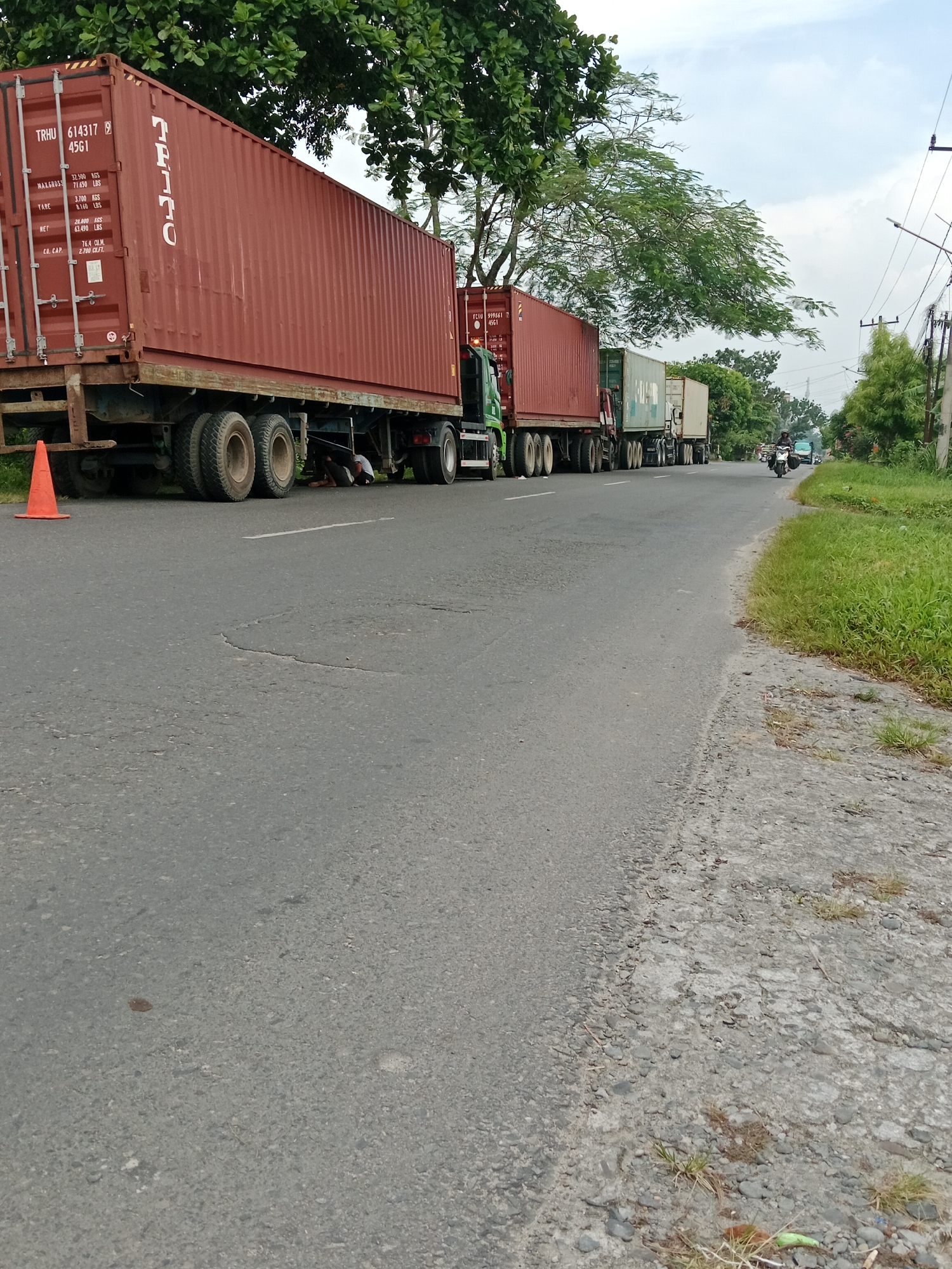 A line of semi-trailers, carrying large cargo containers, is parked on the right side of a two-lane asphalt road. The trailers are deep red, white, and light green. One of the red trailers has text that reads "TRHU 614317 9 4561". Two people are visible under the first red trailer, possibly performing maintenance. An orange traffic cone is placed on the left side of the road, near the front of the first trailer. The road is bordered by green grass and trees on both sides. In the distance, a motorcycle and a blue car are driving on the road. The sky is cloudy. The scene appears to be in a rural or semi-urban area of Binjai, Indonesia, indicated by the tropical foliage and the style of vehicles.