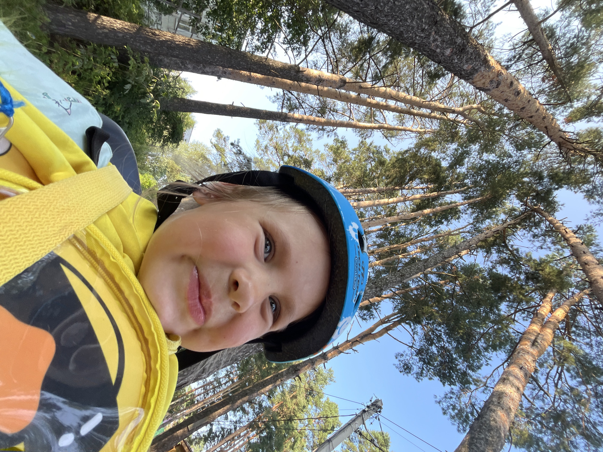 The image shows a close-up, low-angle view of a young child wearing a blue helmet and a yellow hoodie. The child is looking upwards with a cheerful expression. In the background, tall pine trees and a clear blue sky are visible. A utility pole with wires can be seen to the right. The scene appears to be outdoors, likely in a park or wooded area, possibly on the outskirts of Chaikovsky, Russia. The lighting suggests it is daytime, with sunlight filtering through the trees.