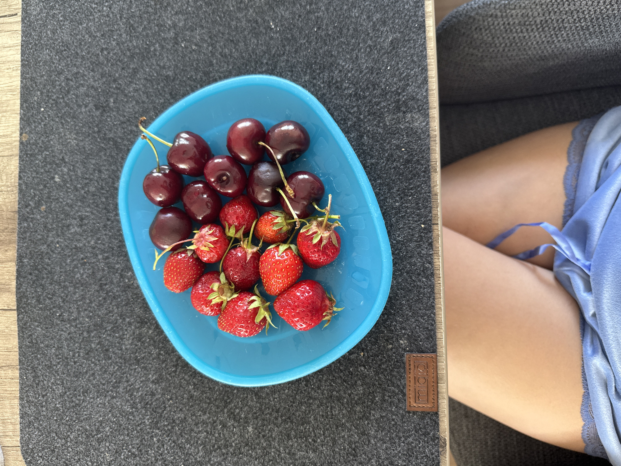 This image depicts an indoor scene in Bruchköbel, Germany, viewed from a slightly elevated perspective. A light blue, shallow, oval-shaped bowl, positioned centrally on a dark gray felt mat, contains a mixture of approximately 10-12 ripe red strawberries with green caps and stems, and 10-12 dark red cherries, some with green/yellow stems attached. The felt mat rests on a light wooden surface. A small, brown rectangular tag, embossed with the word "Miqio", is visible on the mat in the lower-left quadrant. Below the table level, the bare legs of a person are visible, crossed at the knees, wearing light blue undergarments with a lace trim. A portion of a dark gray textured fabric chair is present to the right of the person's legs. The scene is brightly and evenly lit.