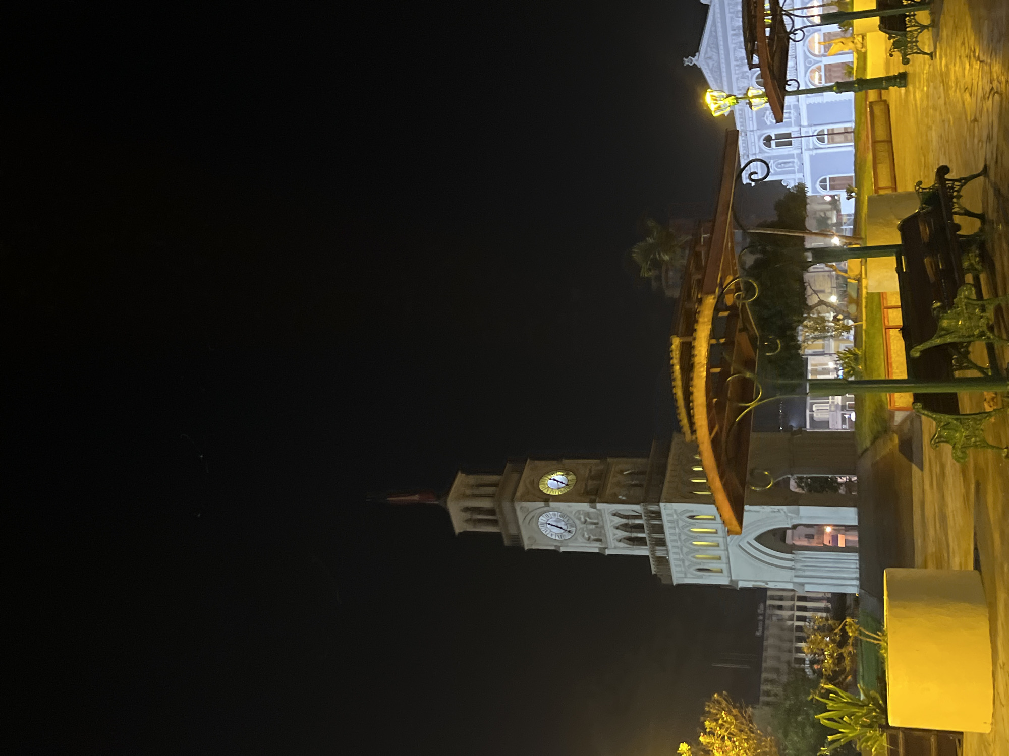 The image shows a night scene in an urban area, potentially in Unknown, Unknown. The most prominent feature is a tall clock tower with two visible clock faces illuminated in the dark. The tower is white or light-colored, and the clock faces are round. In the background, there are other buildings, including one with a blue and white facade and multiple windows. In the foreground, there are elements of street furniture, such as park benches and lamp posts. The scene is illuminated by artificial lights, creating a warm, yellow glow on the objects and surfaces nearby. The overall atmosphere is peaceful and serene, captured at night.