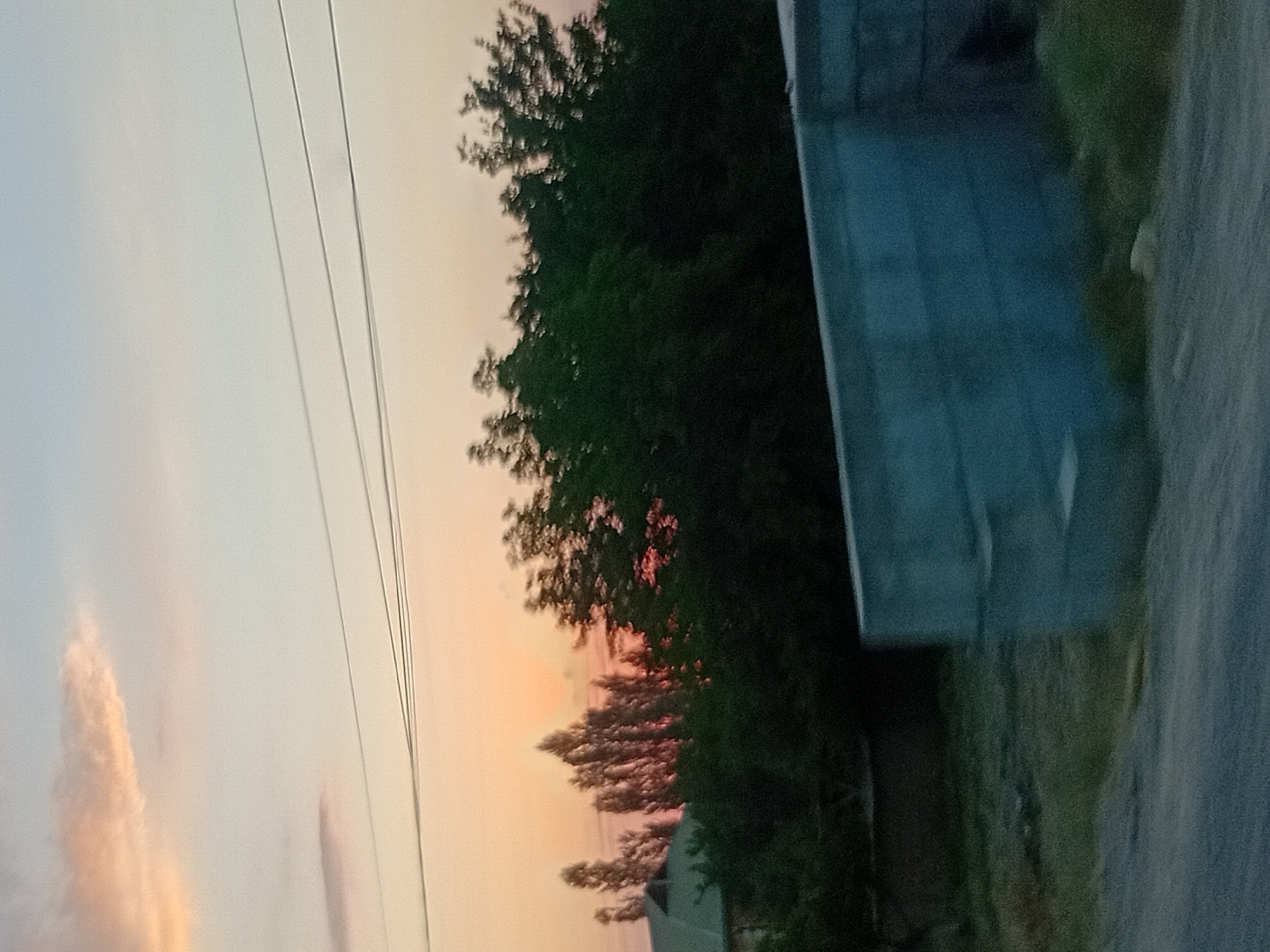 The image depicts a serene sunset scene in an unspecified location. The sky is a canvas of soft hues, transitioning from a pale blue at the top to a warm orange-pink near the horizon. A few wispy clouds drift across the sky, tinged with the colors of the setting sun. Power lines are visible, stretching horizontally across the upper portion of the frame, suggesting an inhabited area.
In the middle ground, a dense line of trees stands silhouetted against the vibrant sky. Some of the trees are tall and slender, their branches reaching upwards, while others are fuller and darker, forming a thick canopy. Behind the trees, the roof of a building with a distinct green hue can be seen peeking through.
In the foreground, a partially constructed concrete barrier runs horizontally across the image. The barrier appears to be made of large, rectangular blocks. The ground in front of the barrier is covered with dry grass and some scattered debris, suggesting a semi-rural or undeveloped area. The overall impression is one of quietude and natural beauty, with the elements of human presence, like the power lines and the construction, subtly integrated into the landscape.
