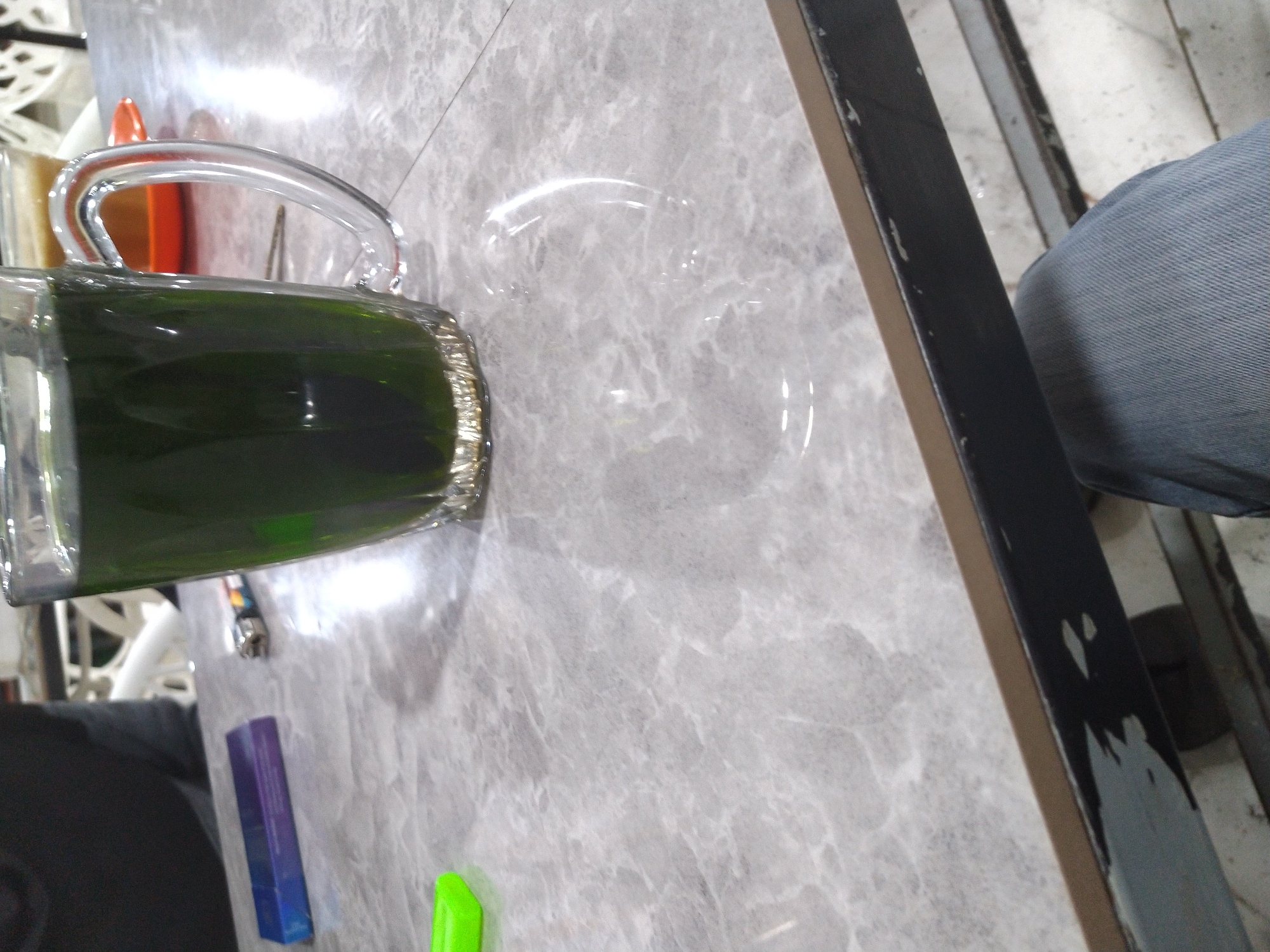 The image captures a close-up, slightly tilted view of a casual indoor setting, likely a cafe, restaurant, or home in Matangkruet, Indonesia. The main focus is on a grey marbled table surface.
On the table, a clear glass pitcher or jug, with a visible handle, holds a substantial amount of dark green liquid, possibly a drink. Next to the pitcher, a clear, empty glass is also visible, indicating a drink might be poured from the jug. Further down on the table, two small, brightly colored objects are present: one is purple and rectangular, possibly a lighter or a small box, and the other is a vivid lime green.
To the right side of the frame, a black table leg or frame is visible, showing some wear and possibly chipped paint. Beyond this, a person's leg, clad in blue jeans, is partially seen, suggesting someone is seated at the table. The background is somewhat blurred but shows glimpses of other elements like white furniture or structures and an orange object, possibly part of the decor. Bright reflections off the table and glass suggest either strong overhead lighting or natural light source. There is no visible text in the image.