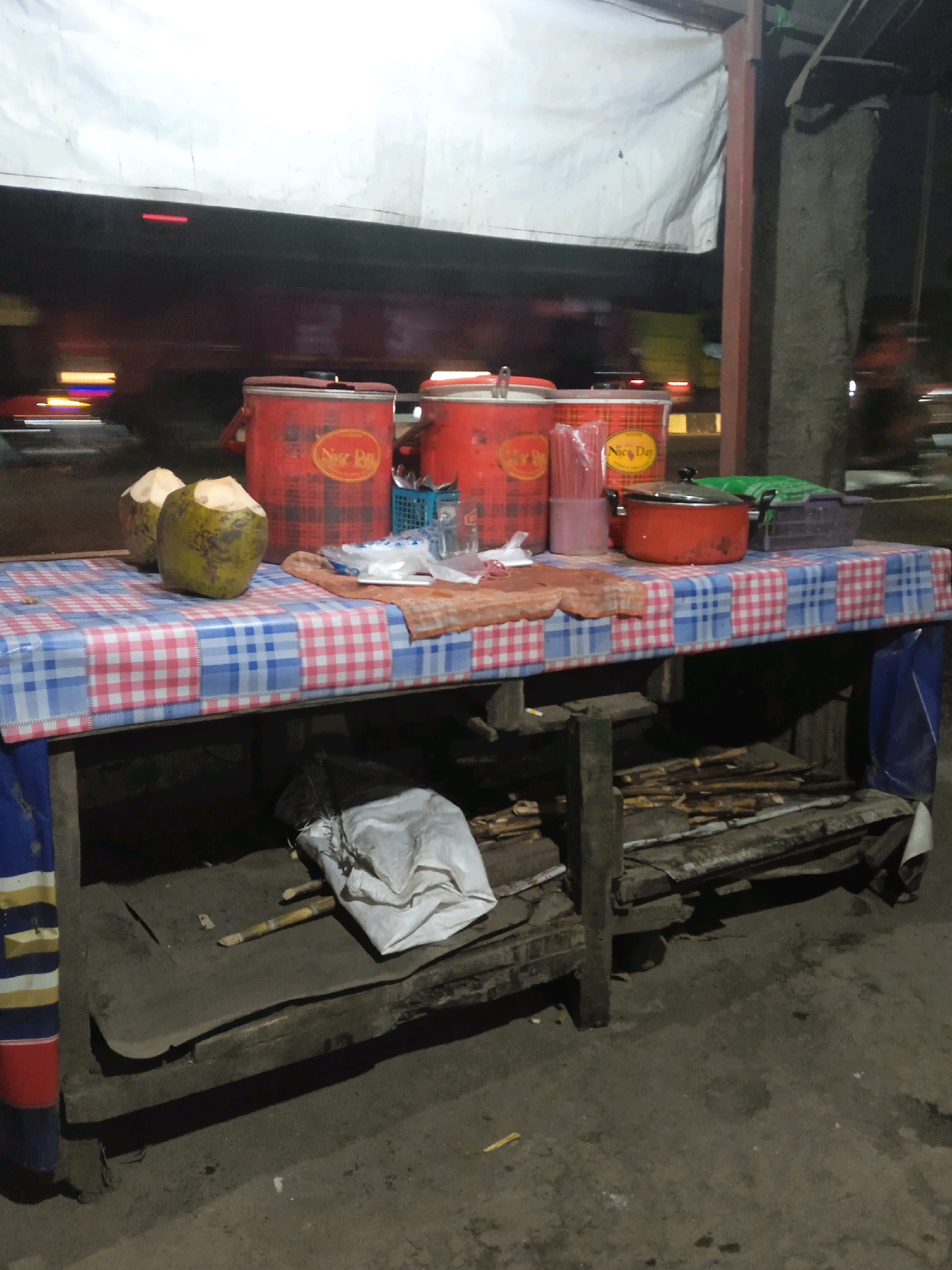 The image depicts a roadside stall at night in Ketapang, Indonesia. A wooden table covered with a red, white, and blue checkered plastic tablecloth is set up. On the table, there are two coconuts, three red cylindrical containers with "Nice Day" branding, a glass of pink liquid with straws, an orange pot with a lid, and a blue plastic basket filled with utensils. A piece of orange fabric and a package of white napkins are also visible on the table. Beneath the table, there is a wooden shelf with pieces of wood and a white bag. The background shows a blurry street scene with passing vehicles, suggesting a busy location. The overall impression is one of a simple, functional street vendor setup.
