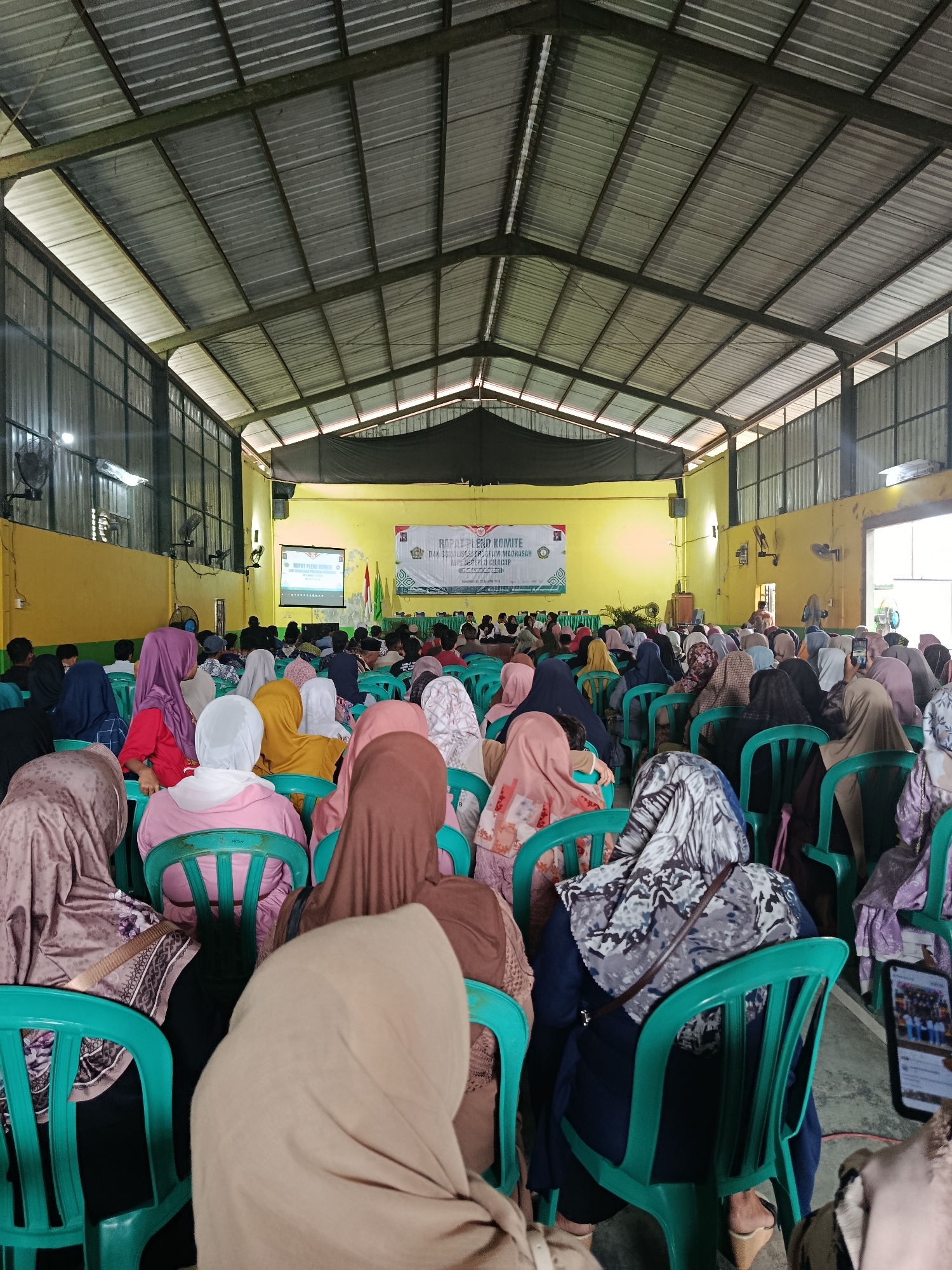 The image depicts an indoor gathering in Kawunganten Lor, Indonesia, likely a public event or meeting. The venue appears to be a large, open hall with a corrugated metal roof supported by exposed beams. The walls are painted yellow, and large windows are visible on the left side. A projector screen displays text and images at the front of the hall, and a banner with text and logos is hung behind it.
A large audience, predominantly women wearing headscarves (hijabs) in various colors and patterns, are seated on green plastic chairs arranged in rows. Some individuals are looking towards the front, while others appear to be engaged in conversation or using their mobile phones. Several fans are mounted on the walls, suggesting a warm climate. The floor is concrete.
The overall impression is of a community event, possibly a forum, seminar, or religious gathering, taking place in a functional, unadorned space. The presence of a projector and banners indicates a formal presentation or discussion is underway.