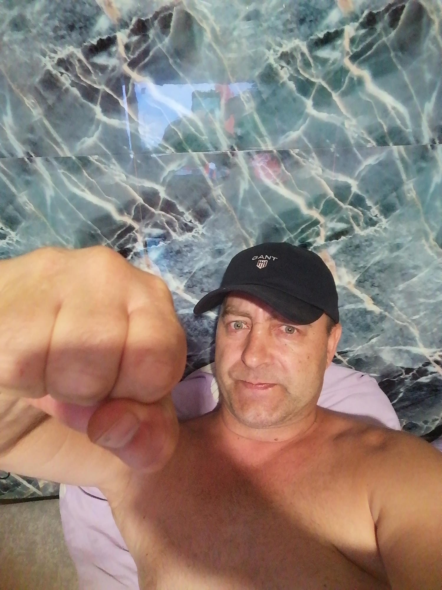 A man wearing a black baseball cap with the Gant logo is shown from the chest up. He is bare-chested and looking at the camera with green eyes. His right fist is in the foreground, obscuring part of the image. He appears to be indoors, with a marbled wall in the background. The image is a selfie.