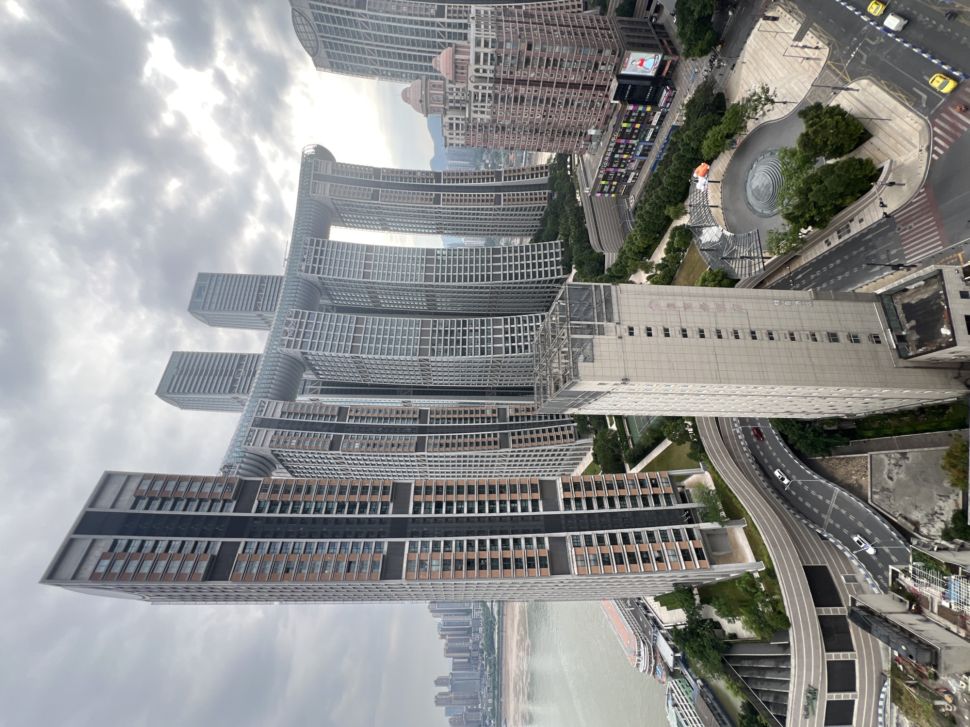 The image displays a striking cityscape dominated by a cluster of modern skyscrapers in Yuzhong District, China. The most prominent structures are the "Crystal" buildings, characterized by their unique design featuring horizontal connecting bridges that create a massive archway. These buildings are tall, with numerous windows and a mix of metallic and reddish-brown facade elements.
In the foreground, a winding road with a few cars, including a white sedan and two yellow taxis, navigates through the urban landscape. The road is bordered by sidewalks and trees. Adjacent to the road, there is a public plaza with a circular water feature and seating areas. A large, light-colored building with many windows stands to the left of the plaza.
Further back, a wide river flows, with several boats docked along the shore, including a large passenger ferry. Across the river, a dense collection of buildings forms the city's skyline, stretching into the distance. The sky is overcast with a mix of dark and bright clouds, suggesting a cloudy or potentially rainy day. The overall impression is one of a dense, modern, and architecturally distinct urban environment.