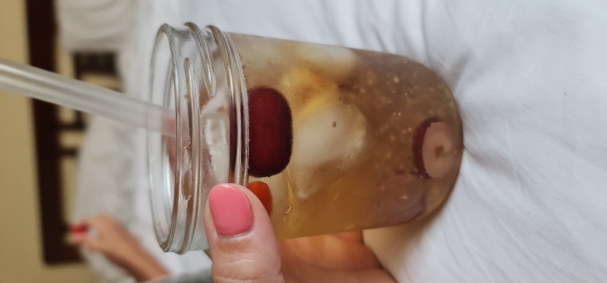 A close-up, eye-level shot depicts a hand holding a glass jar filled with a brown liquid, ice, and sliced fruit, with a clear straw protruding from the top. The jar has threaded ridges near the top, suggesting it may be a repurposed mason jar. The hand holding the jar has a pink manicure on its fingernails. The background includes white sheets or fabric with subtle wrinkles, a blurry figure in the distance, and a glimpse of a wall with decorative elements. The jar is visibly sweating, indicating it is cold. There is no indication of location.
