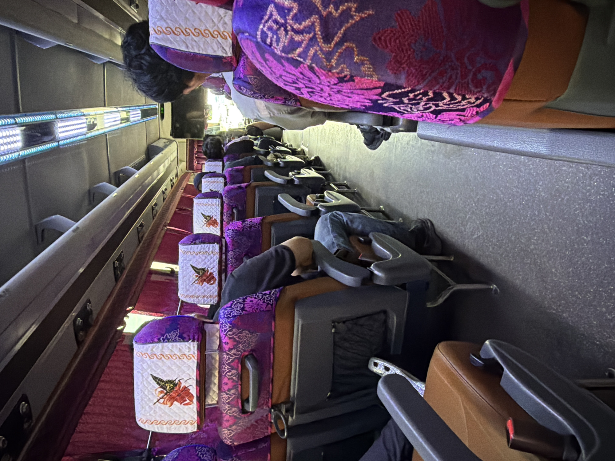 The image shows the interior of a bus with rows of purple and patterned seats. Several passengers are seated, facing forward. The seats have headrests with quilted white covers adorned with an orange and black butterfly design. The aisle is visible, with a textured grey floor. Overhead, fluorescent lights illuminate the cabin, and a television screen is mounted on the ceiling. The overall scene depicts a typical bus interior, likely in transit.