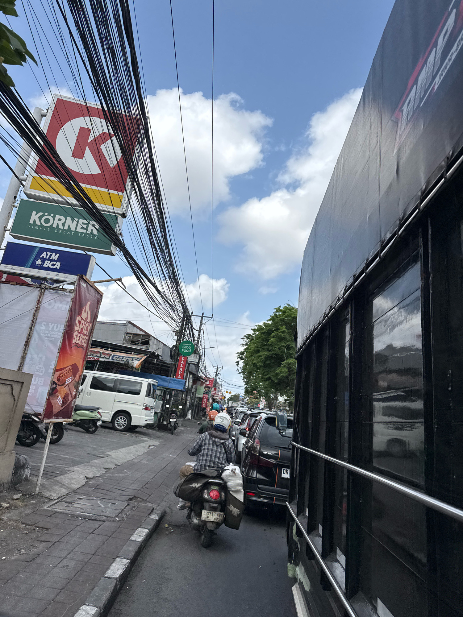 The image captures a street scene in a city, likely in Southeast Asia, given the presence of motorcycles and the style of buildings. In the foreground, a person is riding a scooter, facing away from the camera, carrying bags on the back. The scooter is in a lane of traffic alongside other vehicles, including cars and more scooters. To the right of the traffic lane, a large black truck or trailer with a reflective surface dominates the frame. On the left side of the street, there are several signs, including a red and white "Körner" sign and an "ATM BCA" sign. Buildings line the street, with awnings and various storefronts visible. Overhead, a complex network of electrical wires is strung across the street, a common sight in many urban areas. The sky is partly cloudy with blue patches. The overall impression is a busy, active street with a mix of commercial establishments and vehicular traffic.